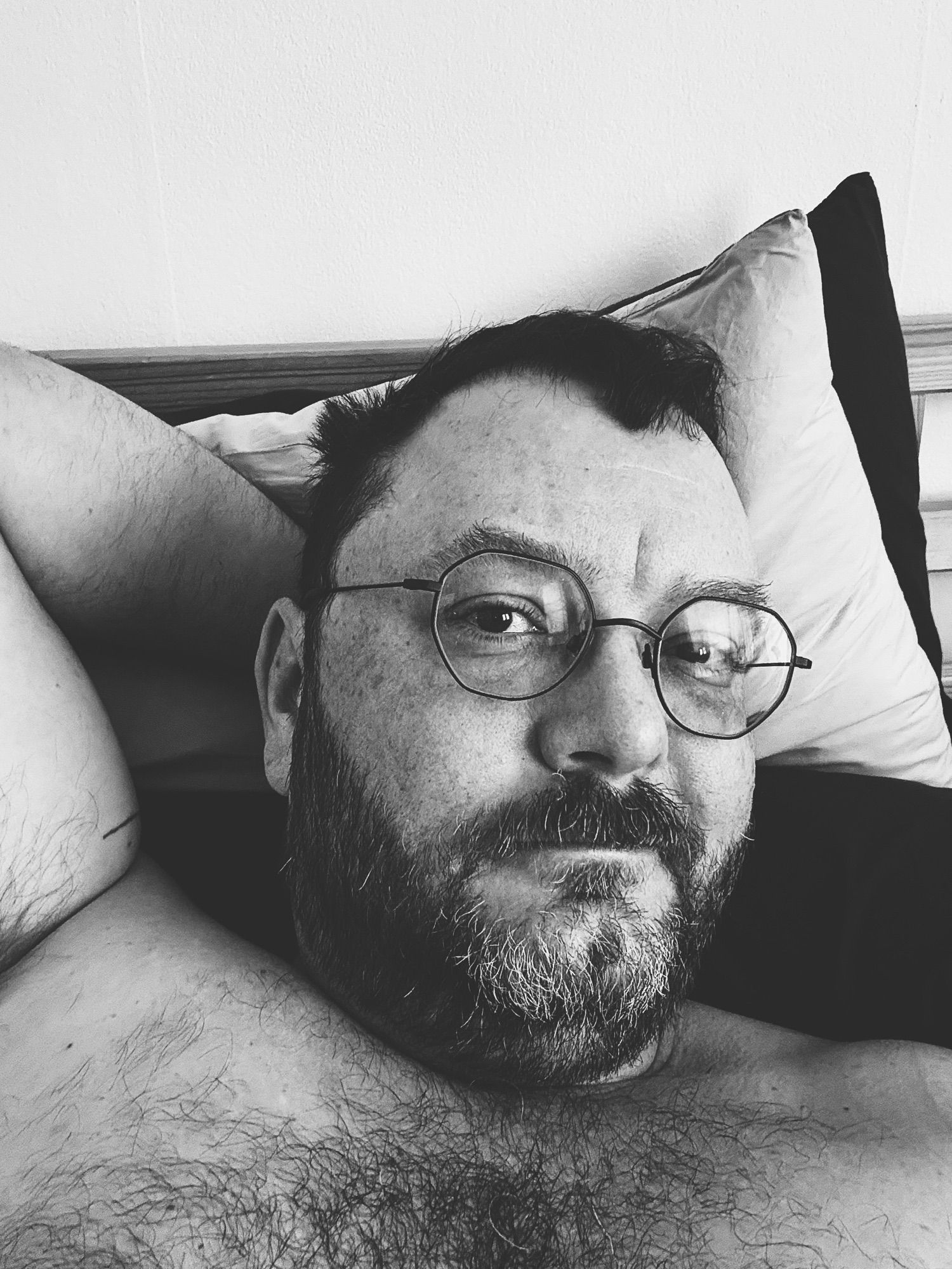 Black and white selfie of me, a middle aged white queer guy, bearded and shirtless, wearing glasses, still lying in bed.