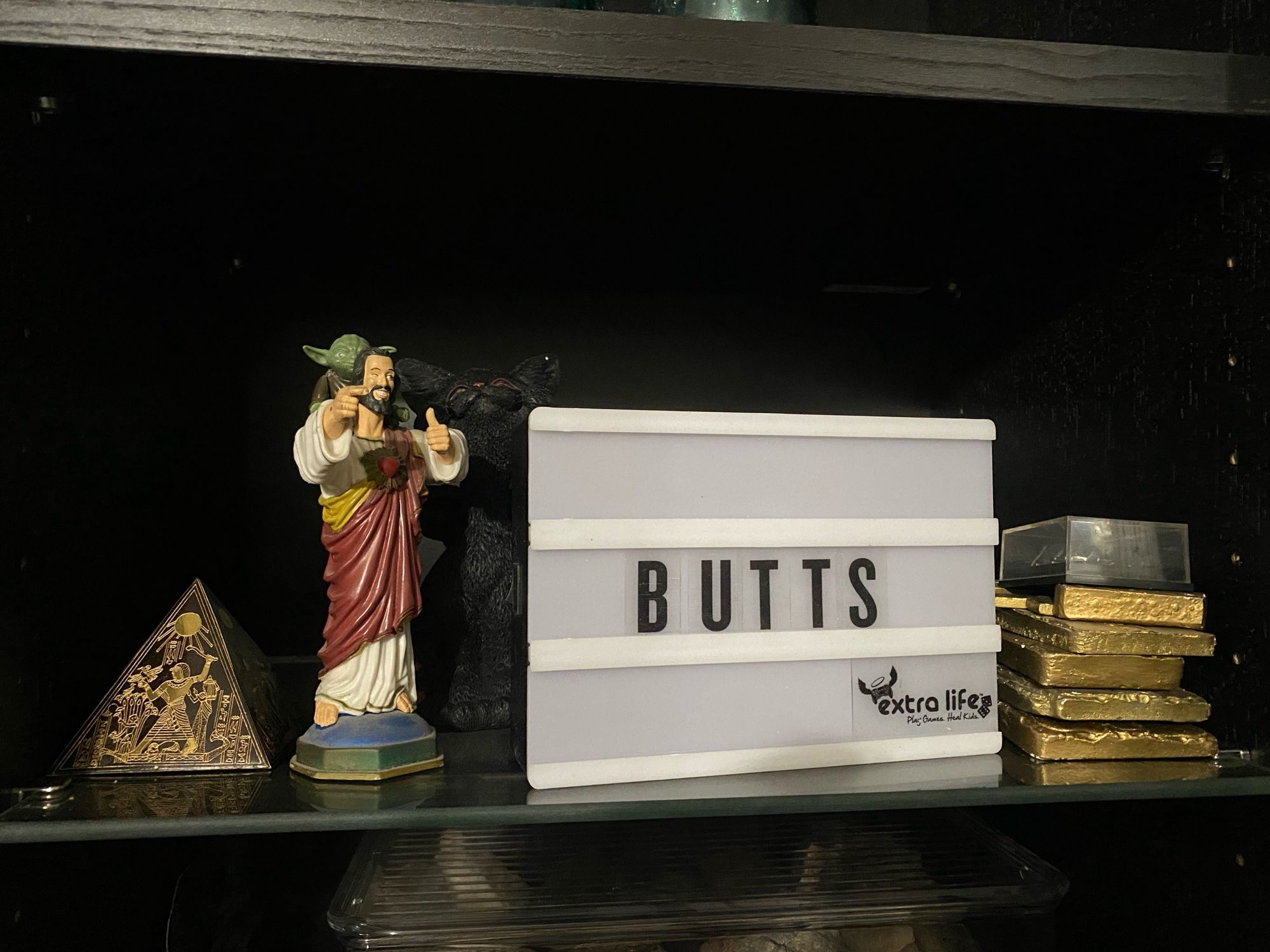 The Buddy Christ giving Yoda a ride, a small pyramid, a stack of gold pressed latinum, and the Extra Life light up marquee with “BUTTS” on it.