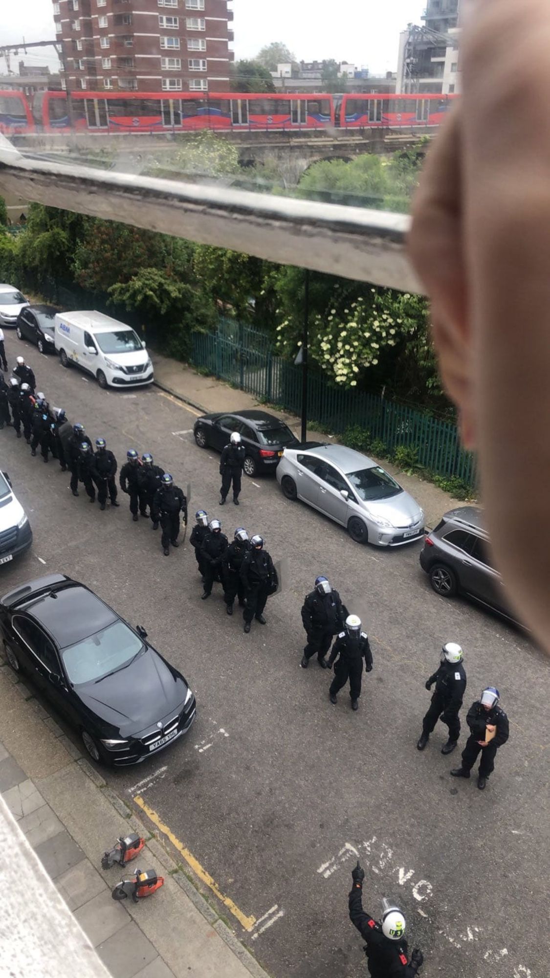 A large number of riot police lined up on a street with a dlr train running behind