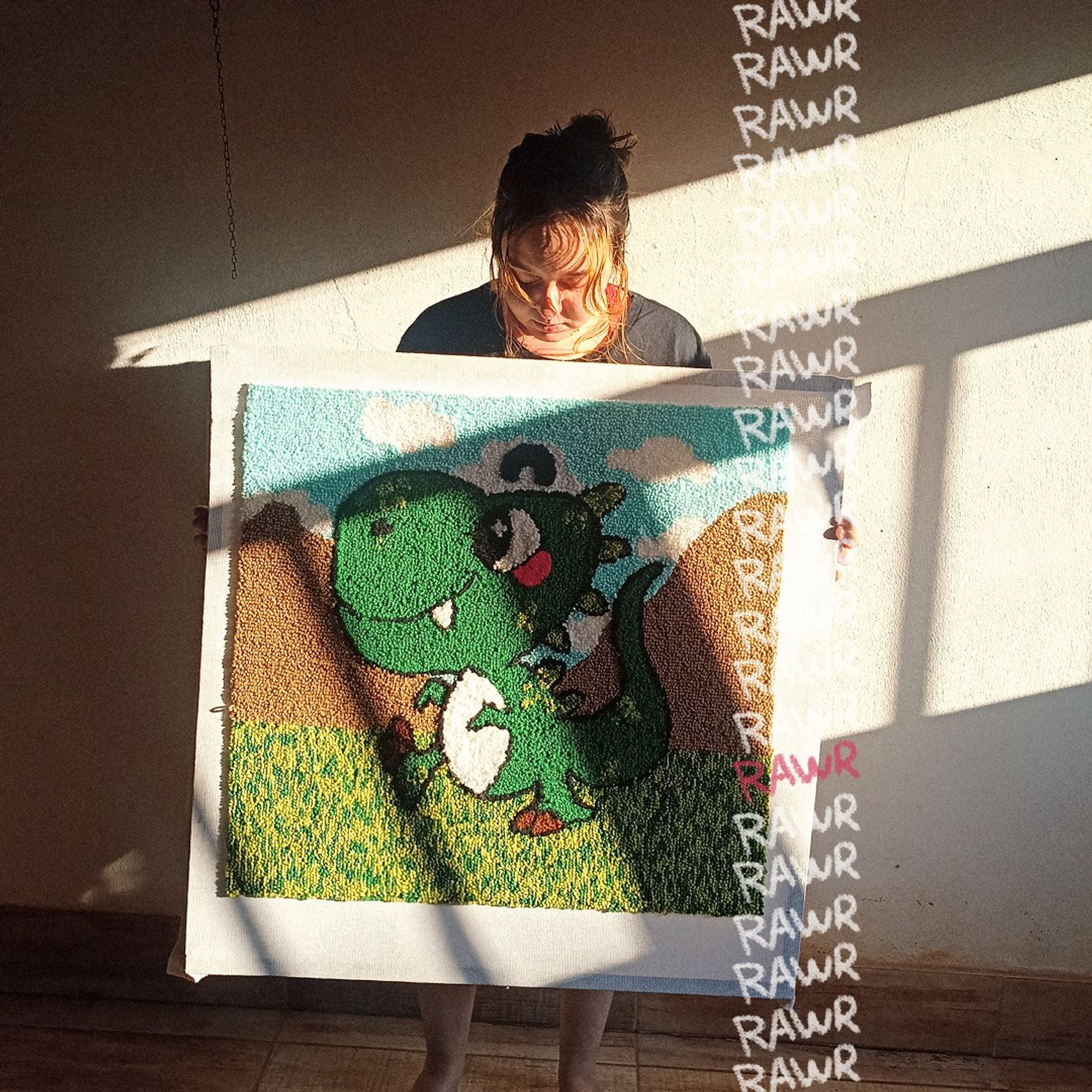 fotografia: a artista giulia gongê segurando uma moldura de 70x70cm de um tapete feito a mão em ponto russo. a imagem principal do tapete é um dinossauro verde estilo cartoon. o dinossauro tem uma expressão alegre, com grandes olhos e um sorriso, e está em um cenário de colinas marrons e um céu azul com nuvens brancas. congê está em pé, olhando para o tapete, com luz solar incidindo lateralmente, projetando sombras na parede atrás dela.