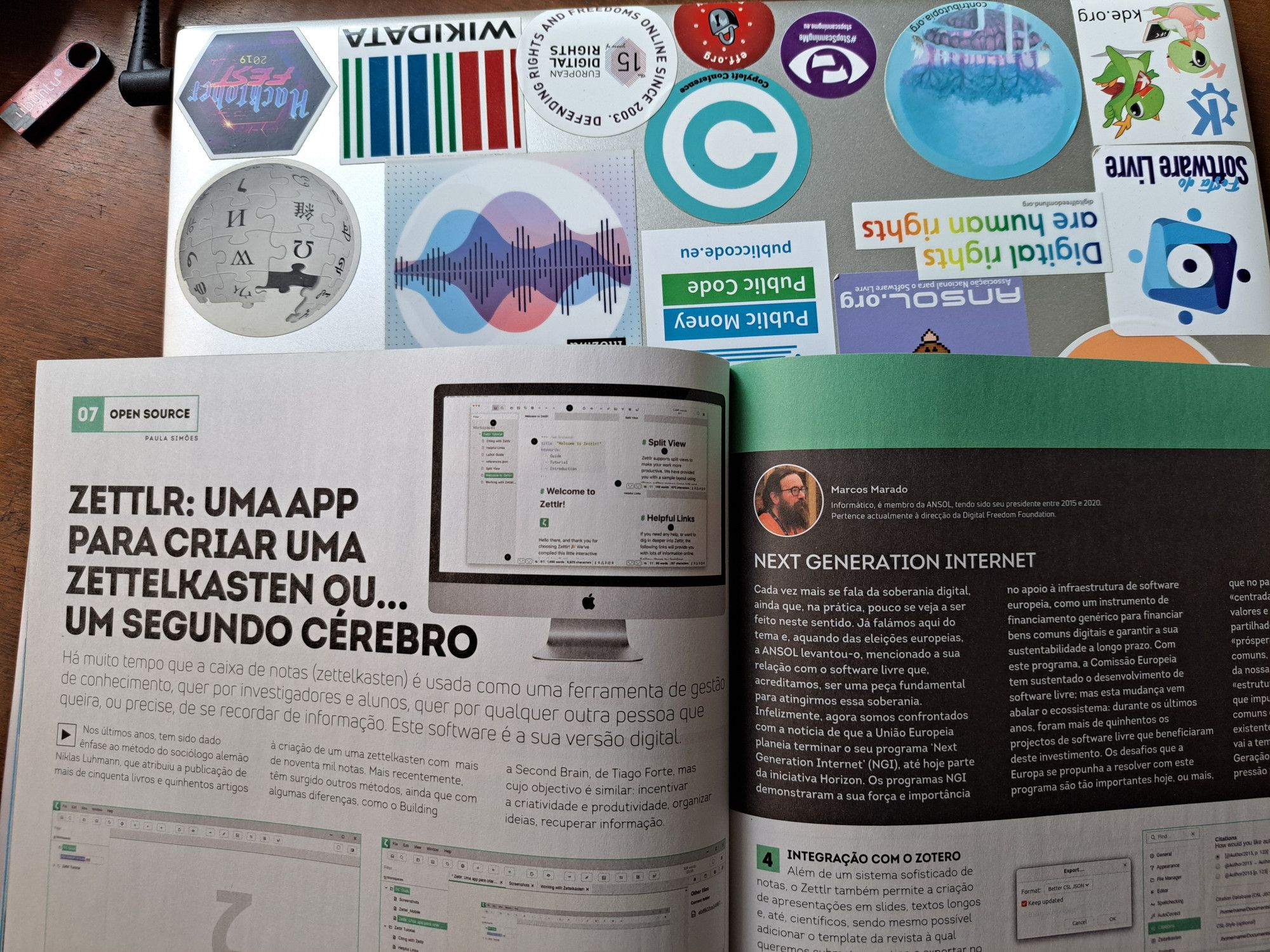 A revista PC Guia em papel aberta nos artigos mencionados sobre um computador portátil com stickers alusivos à cultura e software livres