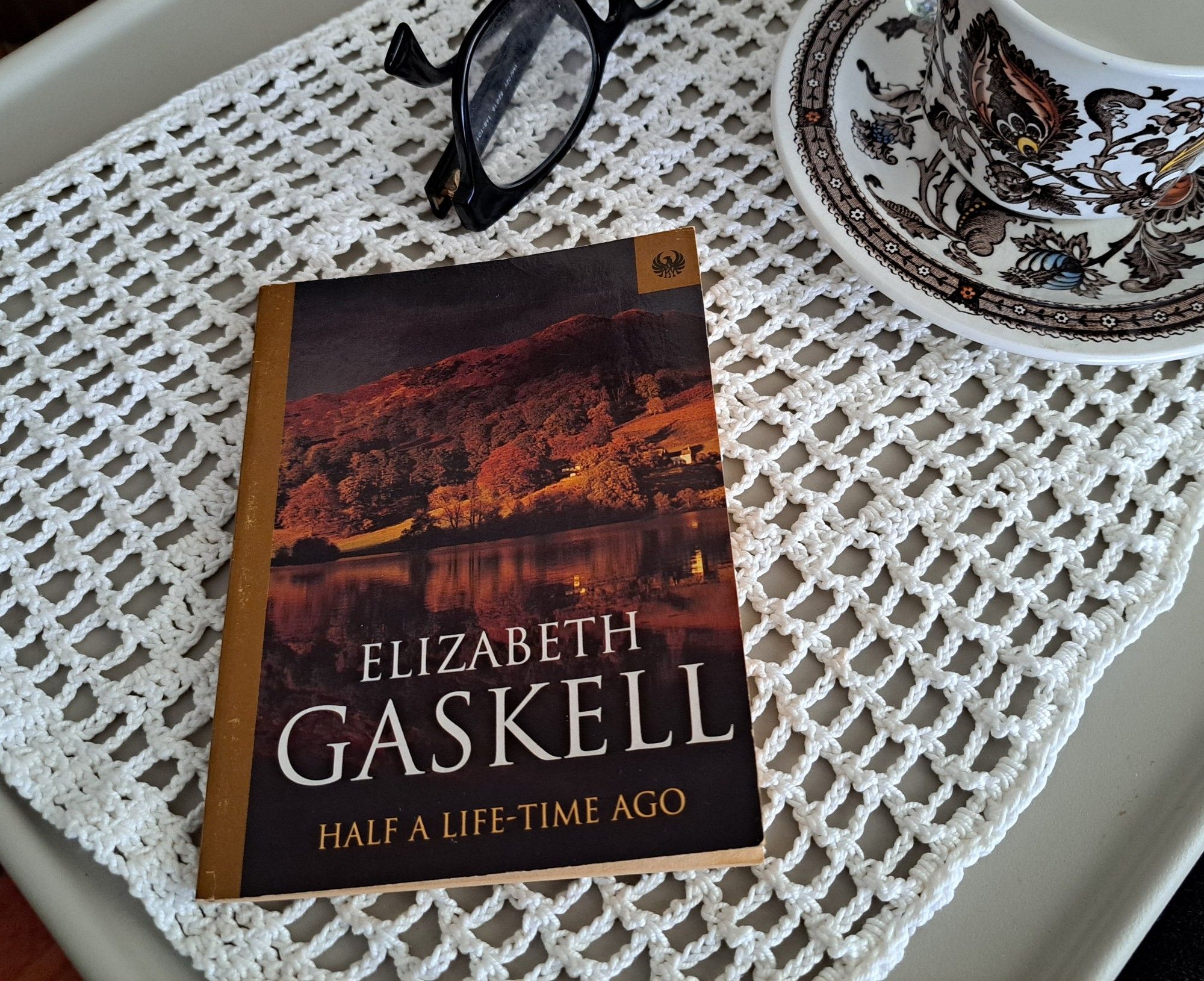 The book Half a time ago by Elizabeth Gaskell, short story with an autumnal colored cover with a tea cup, glasses over a crochet cloth