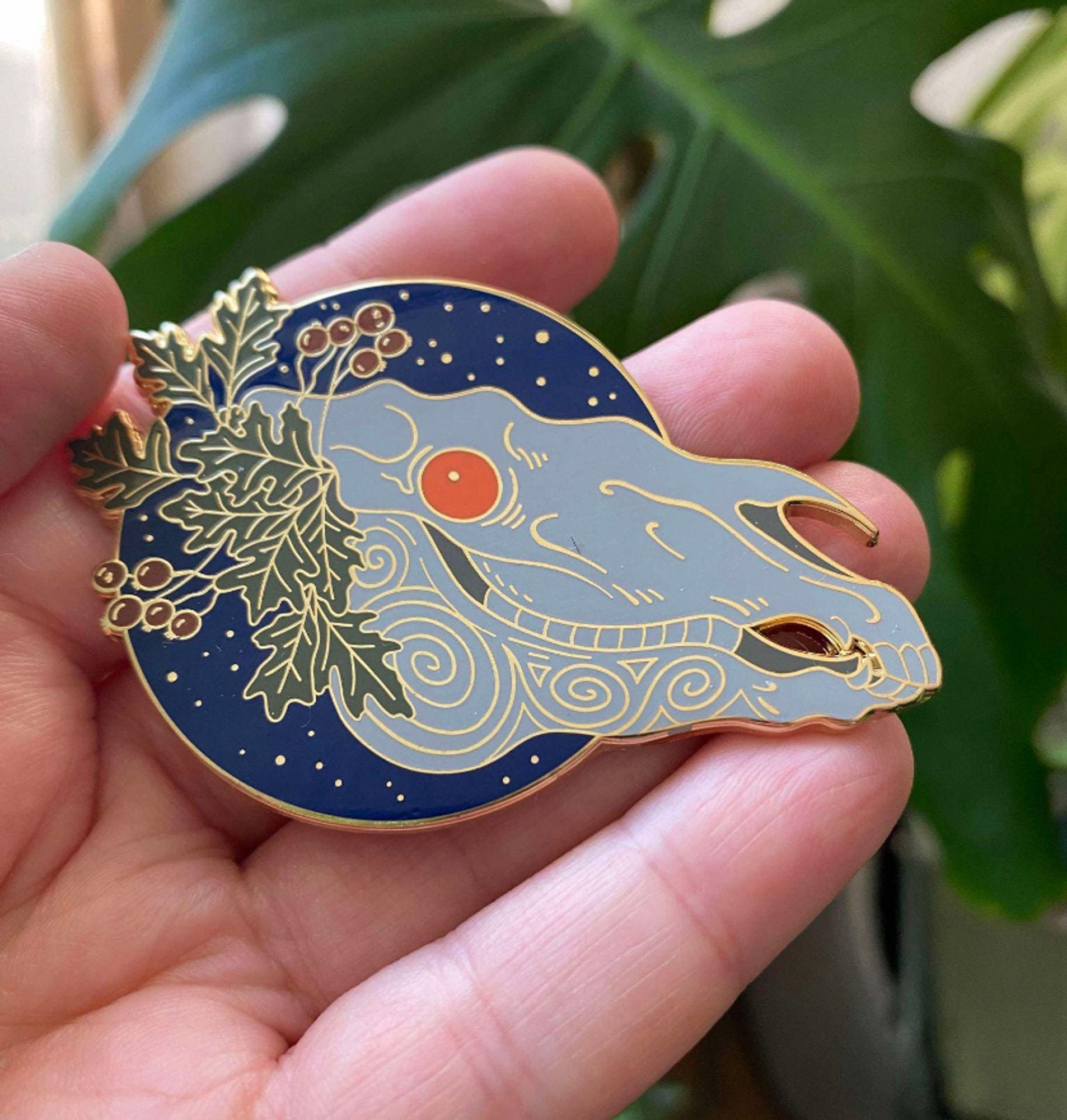 A metal pin badge featuring a white horse skull with bright Orange eyes. It is adorned with Hawthorn leaves and berries and set against a circle of deep blue night sky