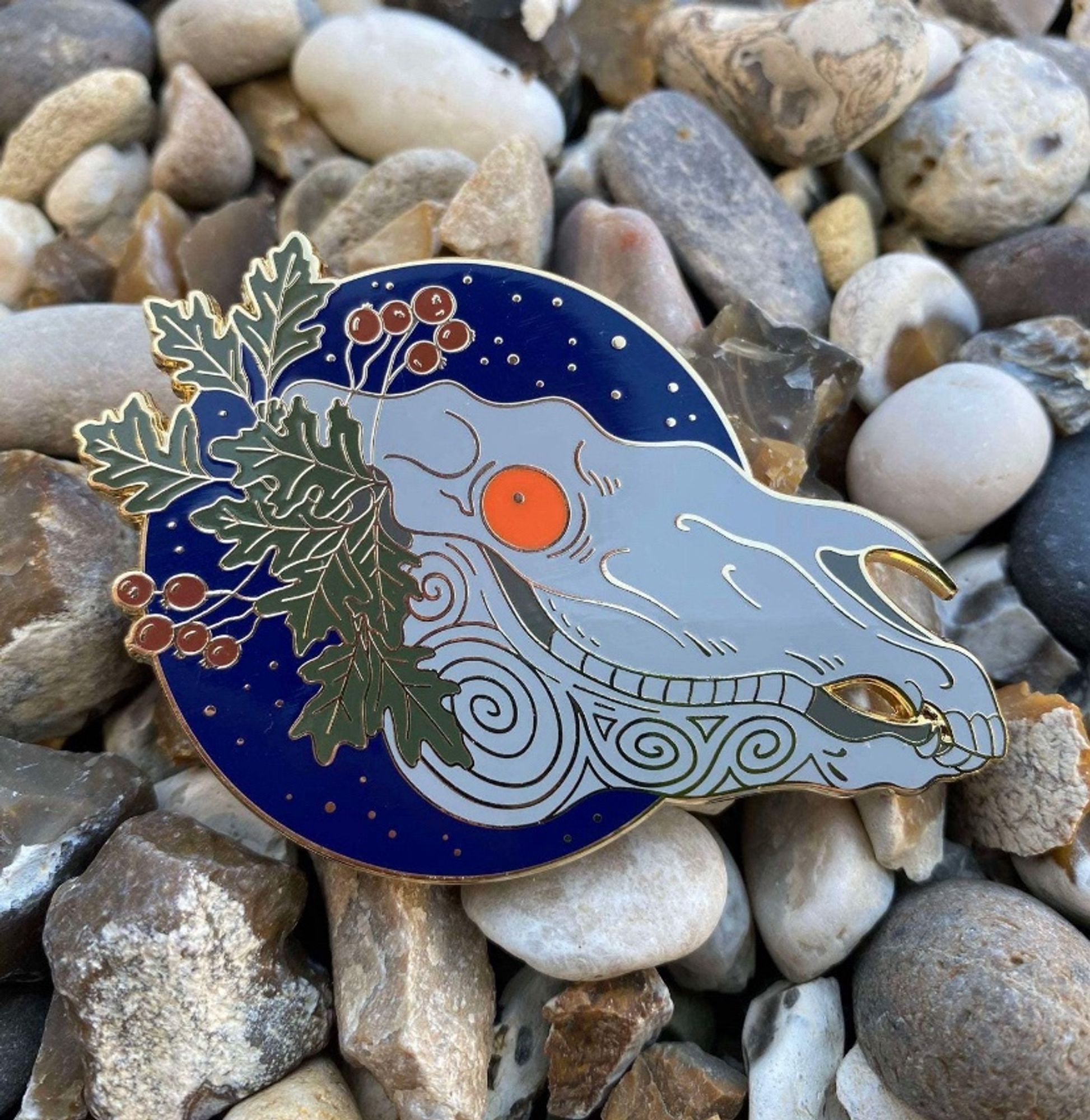 A metal pin badge featuring a white horse skull with bright Orange eyes. It is adorned with Hawthorn leaves and berries and set against a circle of deep blue night sky