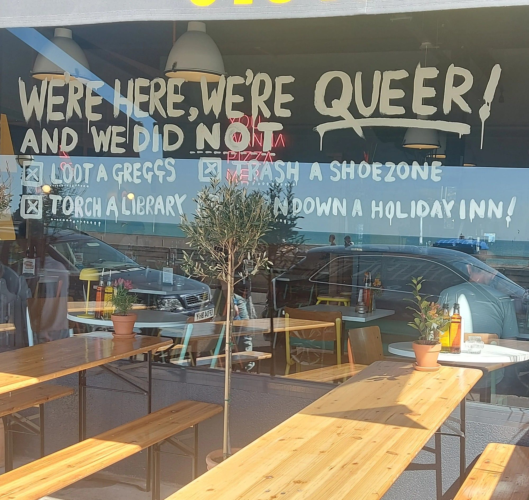 Shop window with sign saying: We're here, we're queer! And we did not: loot a Greggs, trash a Shoezone, torch a library, burn down a Holiday Inn!