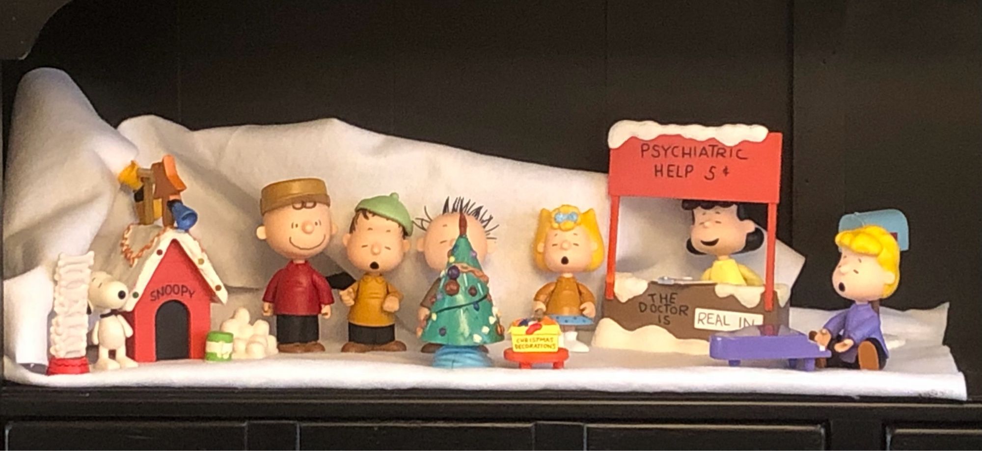 Peanuts figures displayed as Christmas decorations. Snoopy is next to his dog house, which is decorated. Charlie Brown, Linus, Pigpen, Sally, Lucy, and Schroeder are lined up with a Christmas tree in front. Lucy is behind her "Psychiatric Help" stand and Schroeder has his piano.