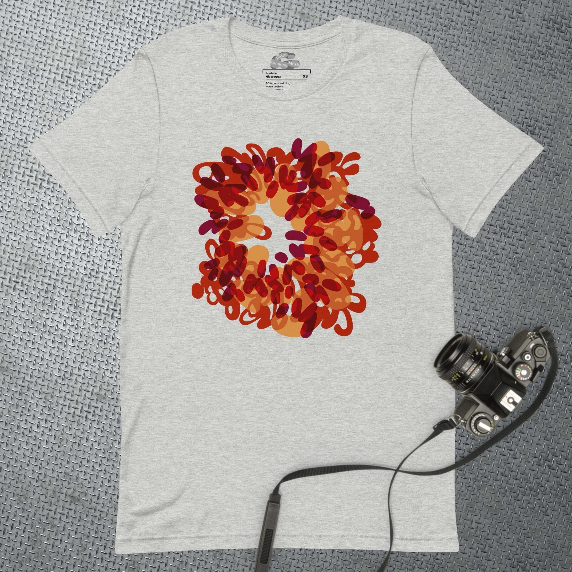 A mock-up of an off-white t-shirt laid out flat on a textured, grey, sheet-metal surface and a black manual camera partially on top of the t-shirt. On the front of the t-shirt is printed an abstracted fire burst with an empty centre, like a wriggly and swirling doughnut of red, orange, and gold yellow overlapping shapes.