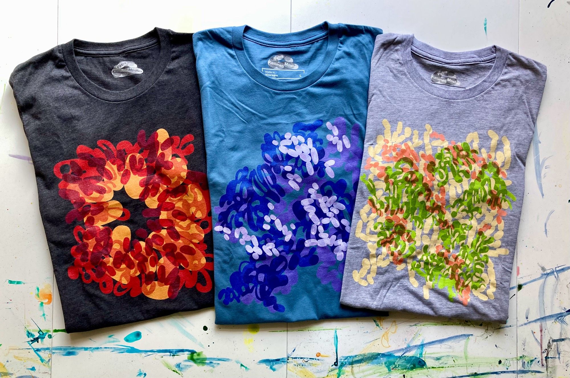 Three folded t-shirts are laid out flat on a white tabletop that has smears and drips of dried paint on it. The t-shirts colours are, from left to right, dark grey heather, a solid true blue, and heather grey. On the front of the t-shirts are printed abstracted, lozenge-heavy designs. The one on the left is a red and orange fire burst, the middle one is a violet and blue foliage design, and the one on the right is a green, pink, and pale yellow foliage design.