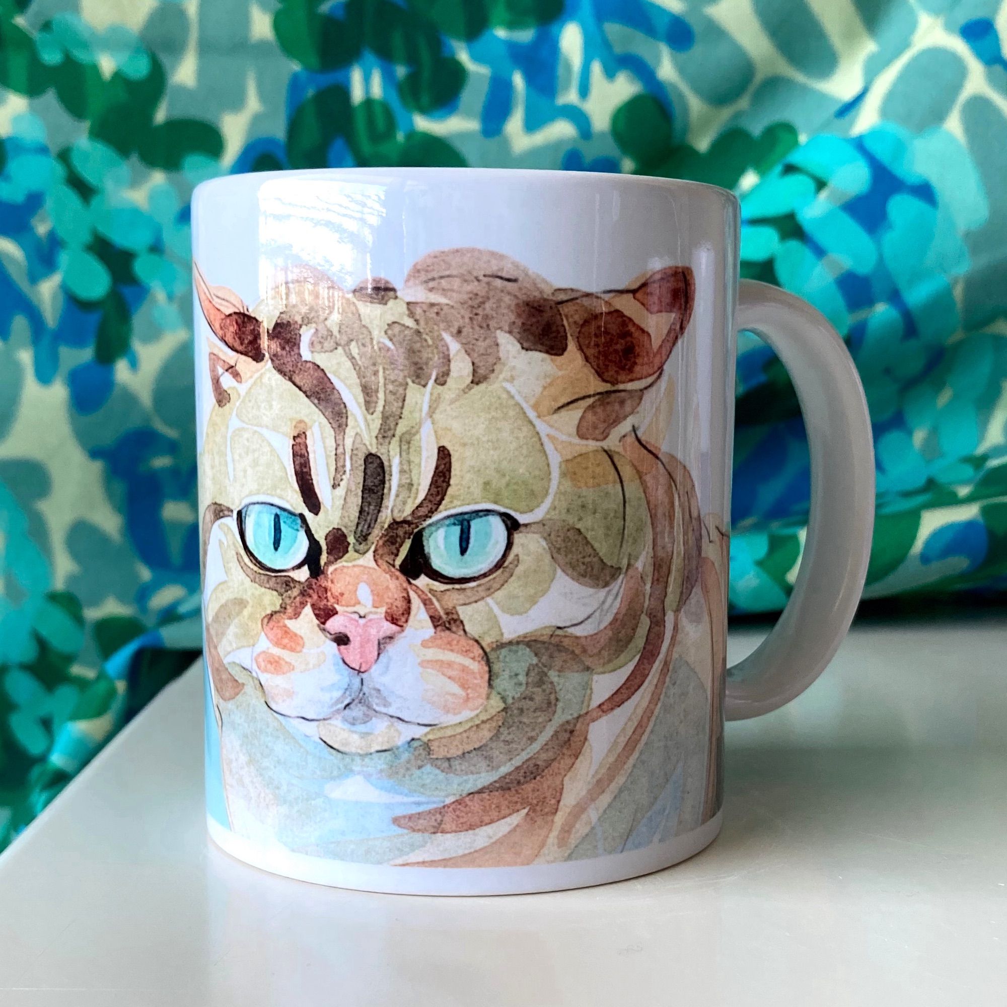 A white, glossy mug with its handle to the right. Printed on the mug is a cat's face with green eyes staring out at the viewer, its ears in airplane mode. It is a watercolour illustration digitally printed on the mug.