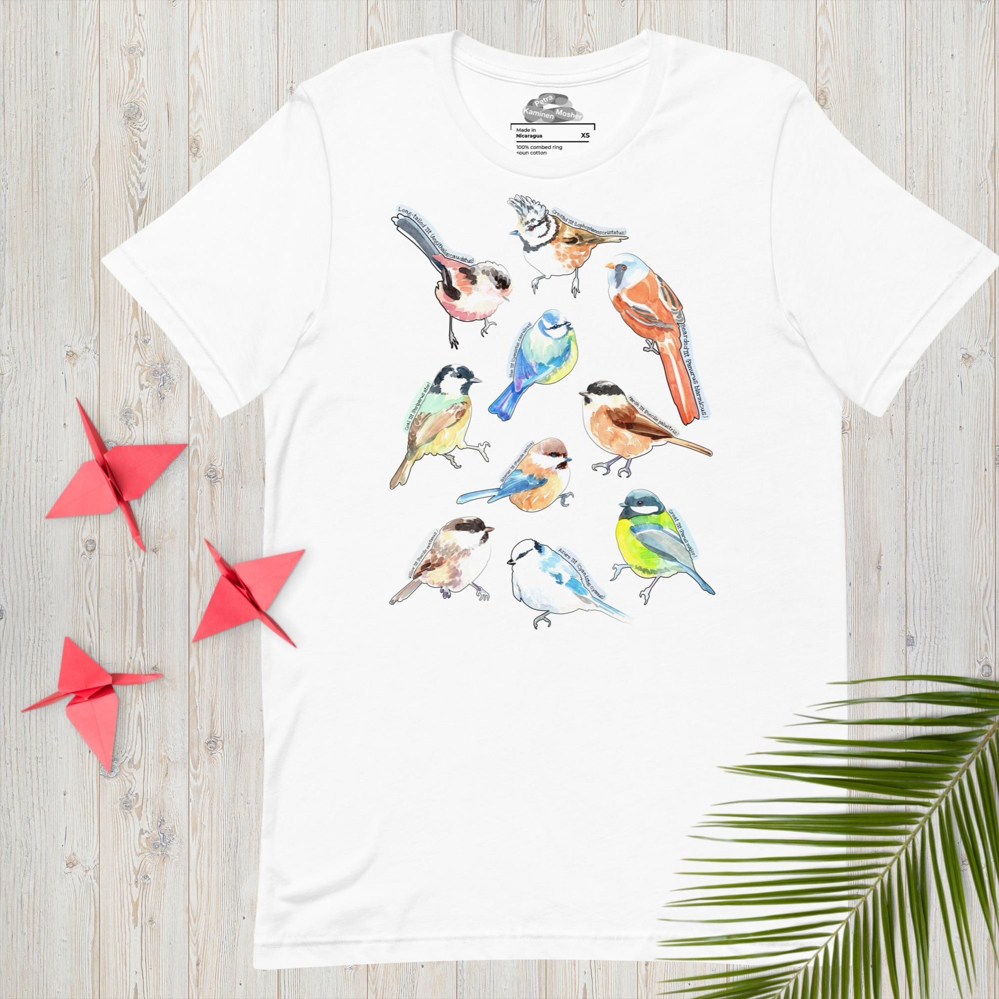 A mock-up of a white t-shirt laid out flat on a whitened wood floor with a palm frond and three red origami cranes placed on top of it (in an artistic manner). The t-shirt has a group of watercolour bird illustrations on the centre front of it. There are ten birds, all known as "tits" in their common English names. The birds have their common and Latin names printed along their top line or tail. The birds are: the Long-tailed tit, Crested tit, Bearded tit, Blue tit, Coal tit, Marsh tit, Siberian tit, Willow tit, Azure tit, and, last but not least, Great tit.