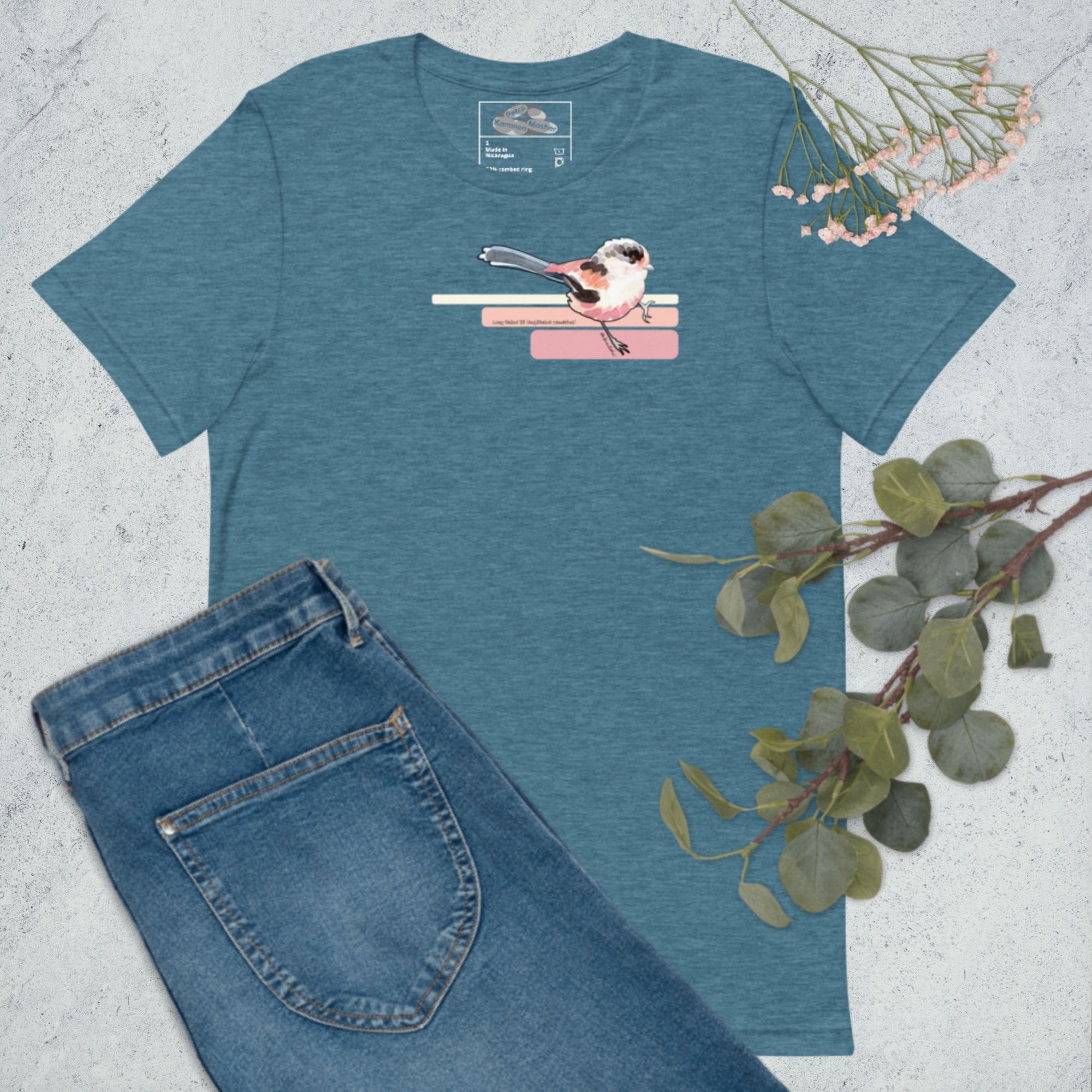 A mock-up of a dark heather teal, short-sleeved t-shirt laid out on a grey marble floor with sprigs of tiny pink flowers, a branch of green leaves, and a pair of dark blue denim jeans overlaid on it. The t-shirt has a medium-sized watercolour illustration of a long-tailed tit on the upper right side of the front of the shirt. The bird looks like it is sitting on three elongated bars of colour similar to the bird.