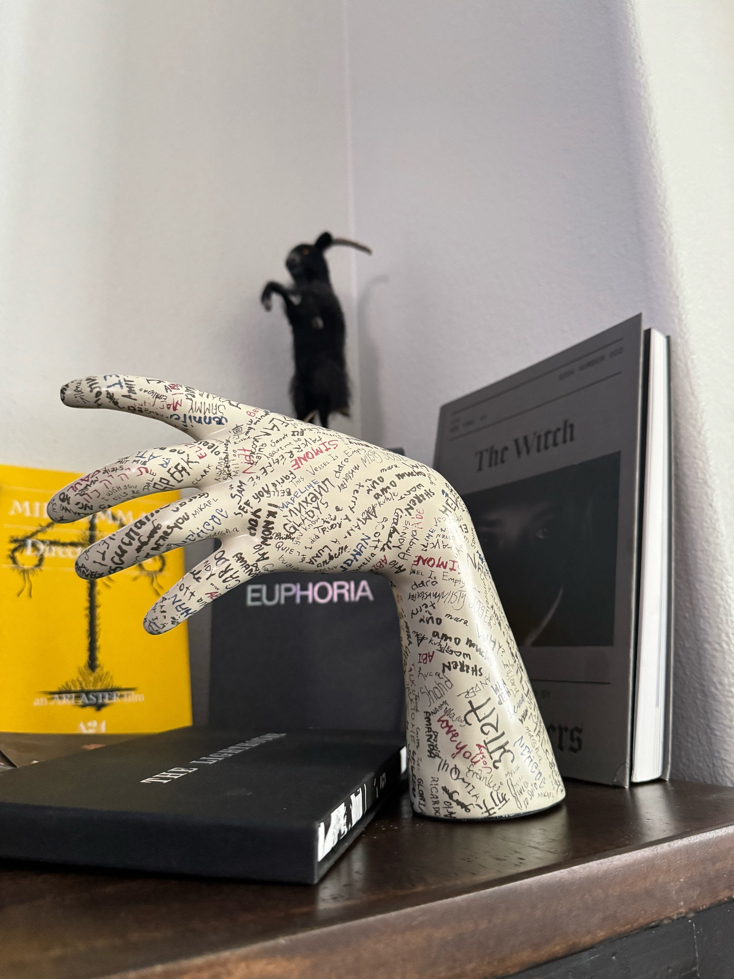 The image shows a display featuring a hand sculpture from the A24 film Talk to Me, covered in handwritten text, along with a few other objects. In the background, there is a dark, standing figure of a goat that seems inspired by Black Phillip from The Witch. A yellow book or DVD case for Midsommar is visible on the left, with a Euphoria box placed between that and a book that says The Witch, referencing the movie of the same name. The overall scene gives off a distinct A24 vibe.