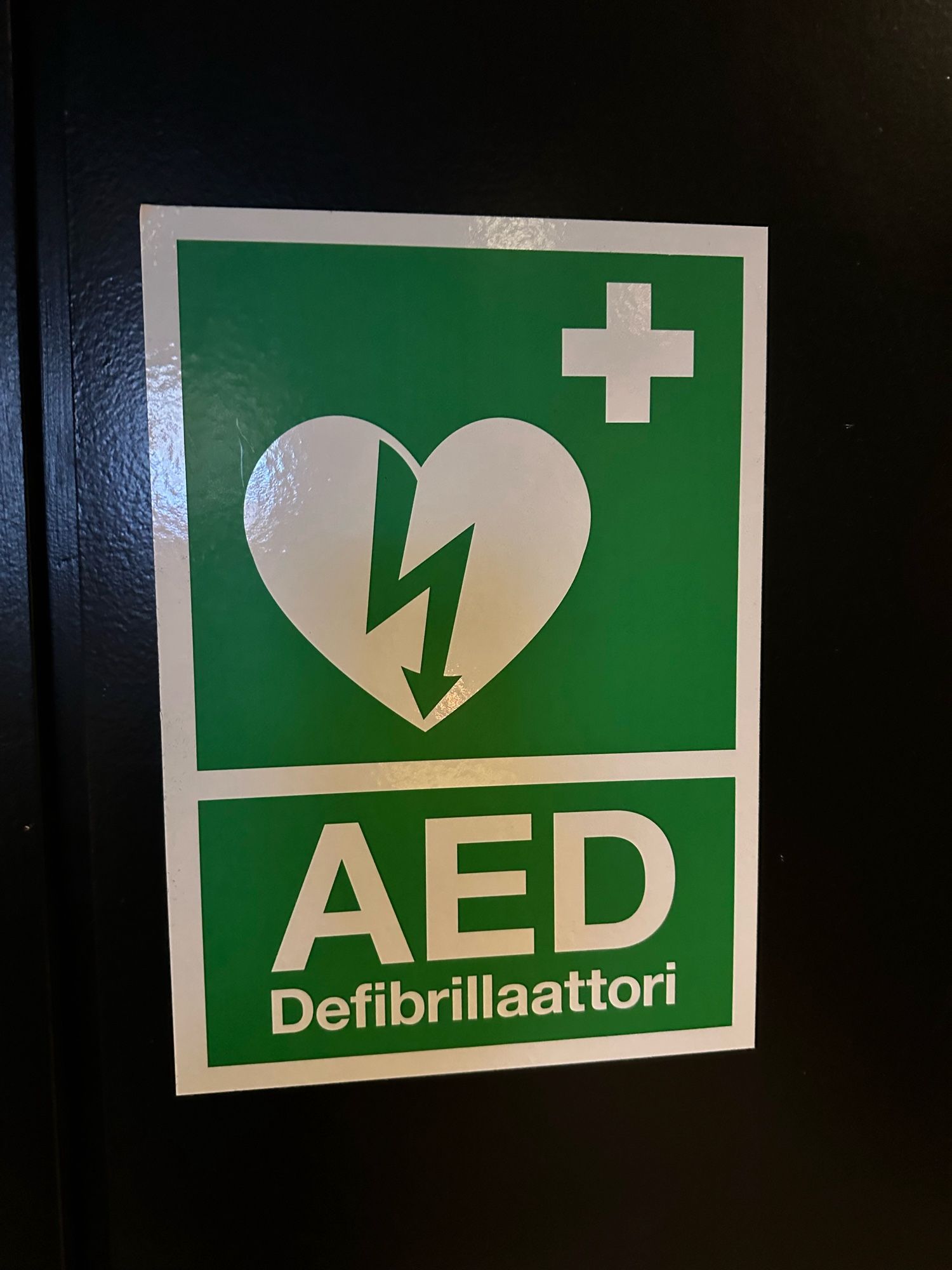 Symbolbild „AED“ 
Hier auf finnisch „Defibrillattori“. 
Die Hinweis-Bilder sind aber überwiegend identisch in vielen Ländern. 

Man sieht einen grünen Hintergrund und ein weißes Kreuz rechts oben. In der Mitte ist ein Herz mit einem Pfeil nach unten (für Strom) darunter die Buchstaben AED und 
Defibrillattori.