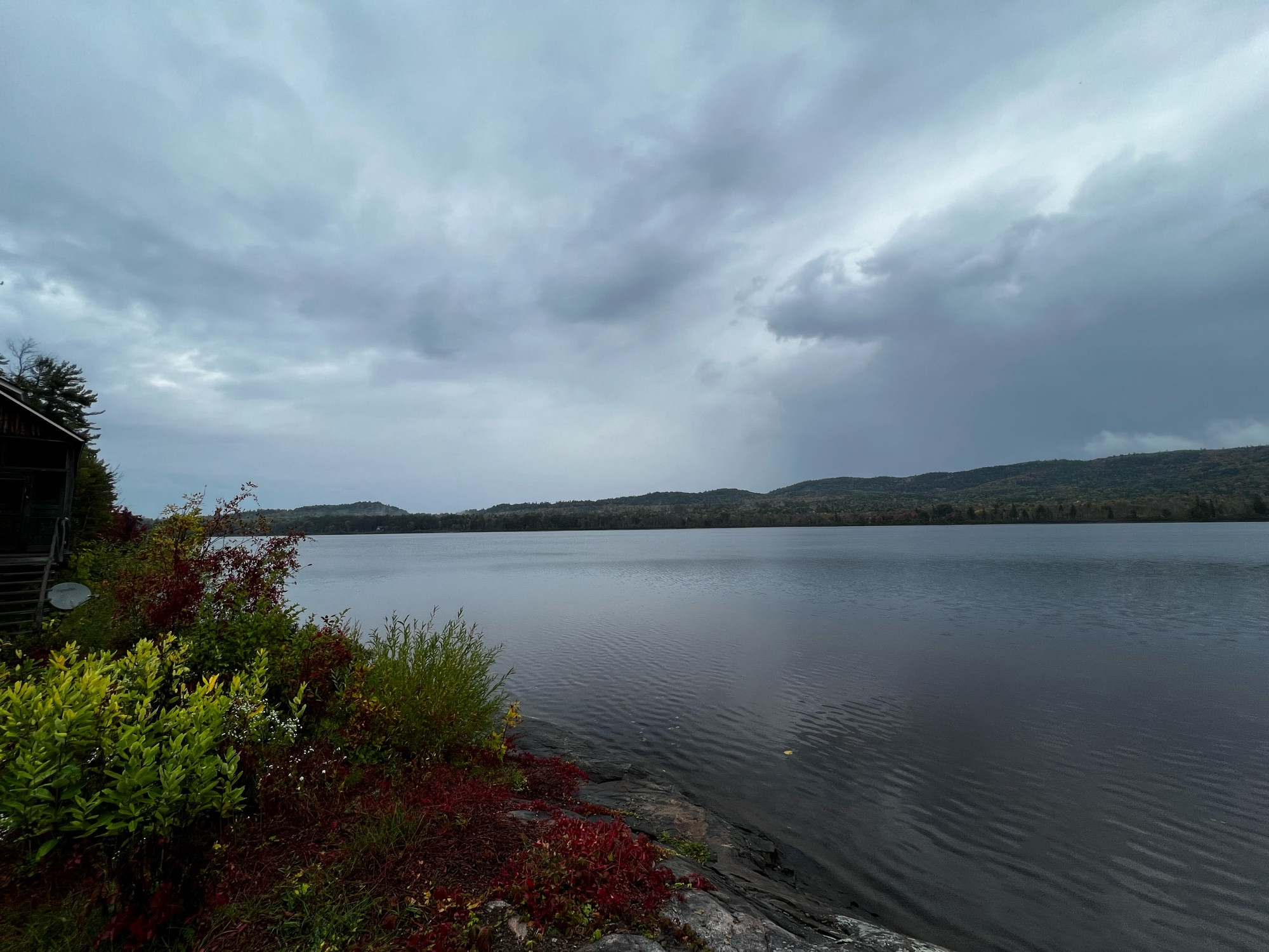 View of a lake on an overcast day. Red and green vegetation line the nearby shore and small, rolling mountains can be seen across the way.