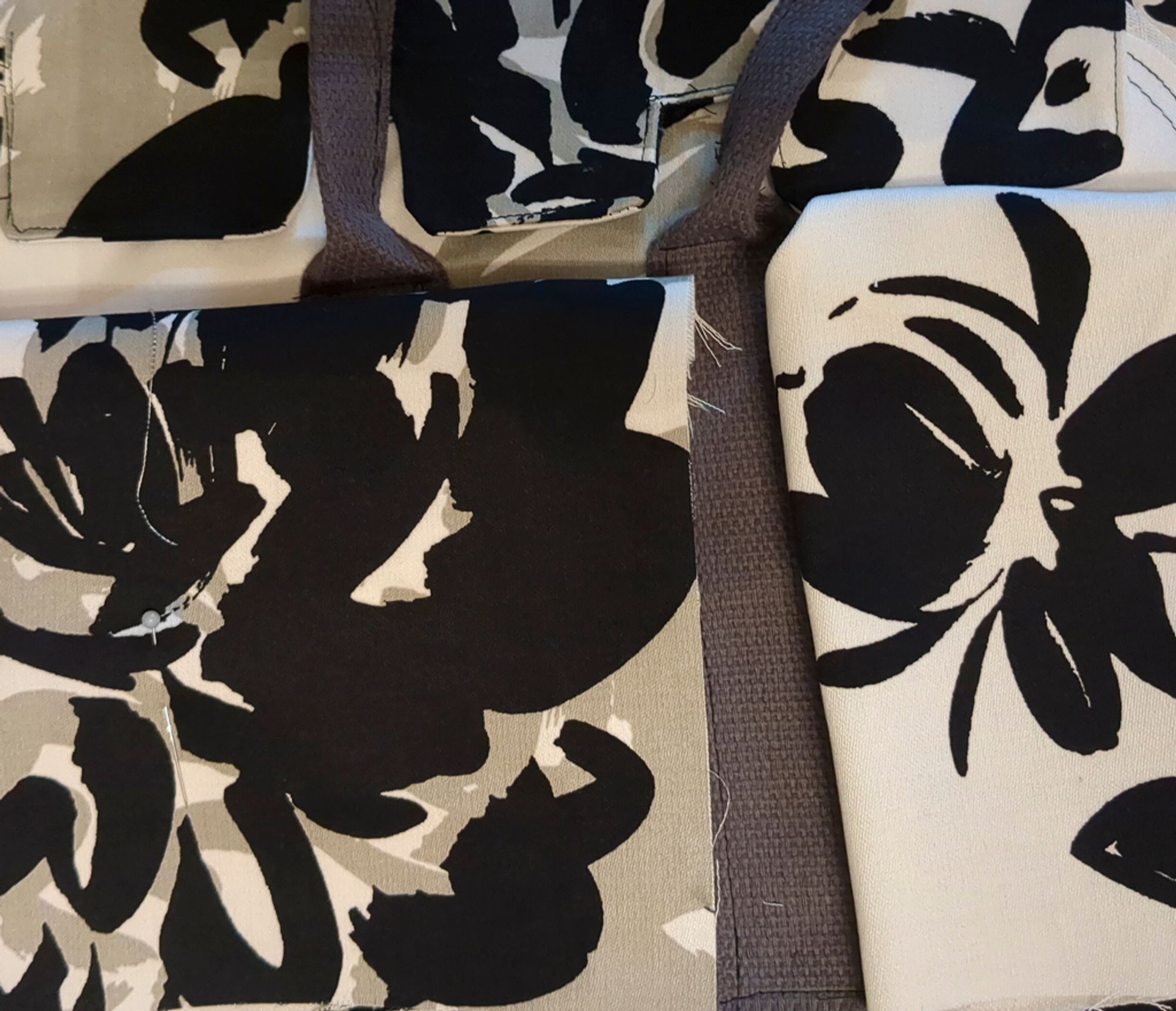 A partially assembled handbag with dark grey cotton strapping, and a white twill with huge splashy black and grey flowers. One pocket piece is mostly white with a smaller black flower. The rest of the pieces are mostly black and grey.