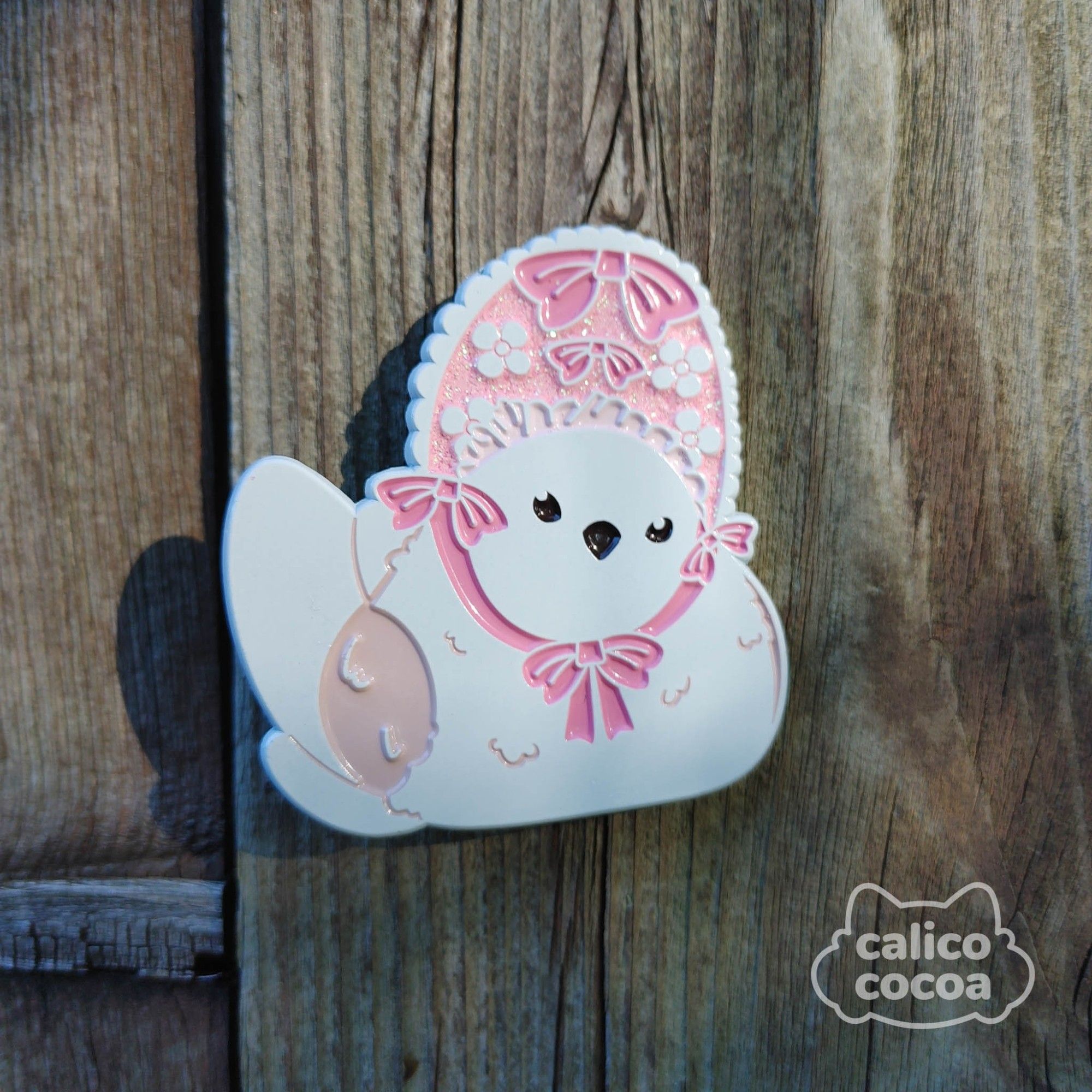 A soft enamel pin dyed white. It features a small white bird wearing a pink bonet with white flowers and ribbons.