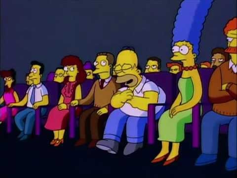 Still from the television show the Simpsons showing Homer sitting next to Marge in a theater surrounded by townspeople. Homer is laughing hard and some of the townspeople are glaring at him.