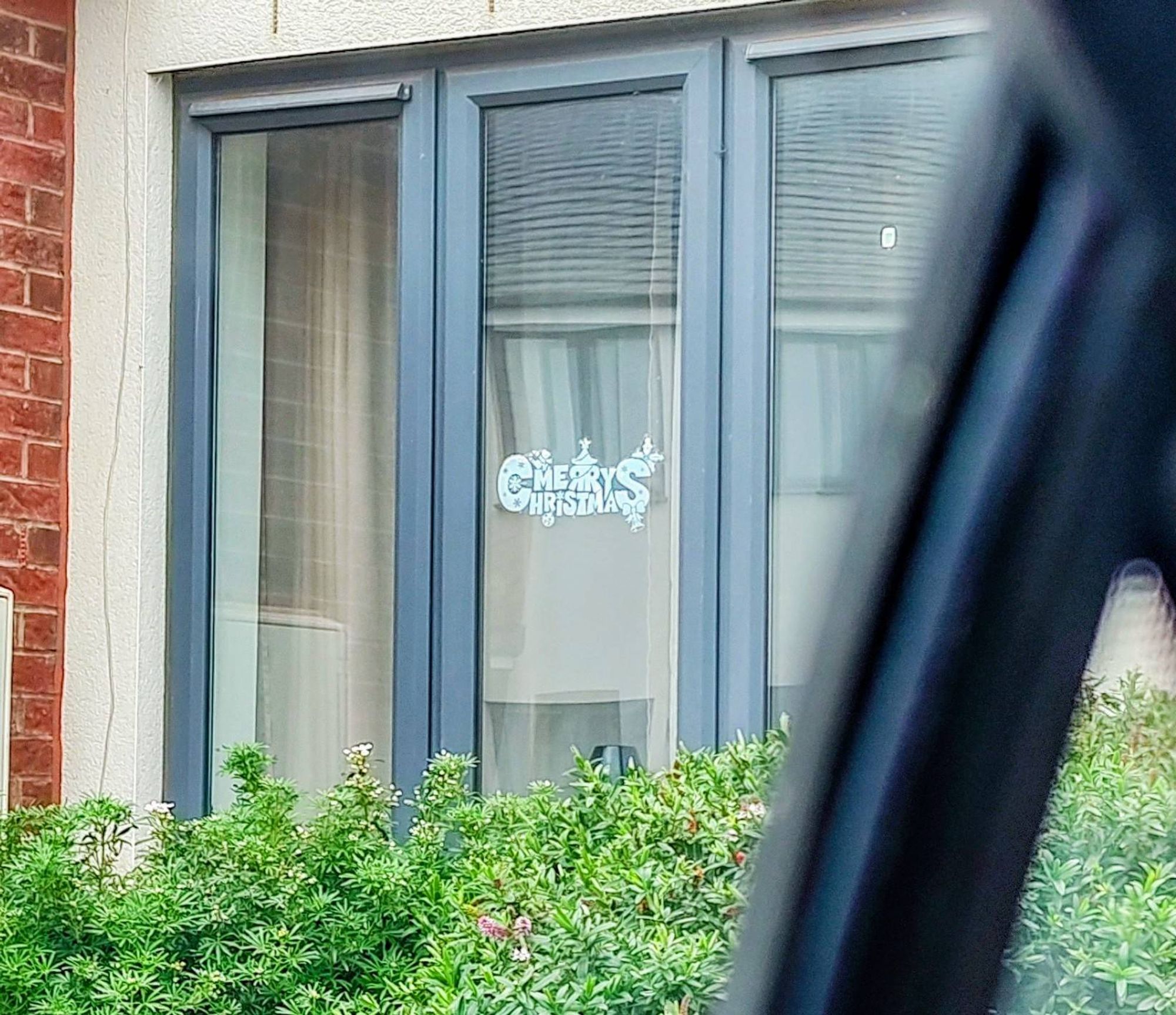 The image shows a window with "MERRY CHRISTMAS" written on it in white. The window is part of a house with brick walls and a gray frame. There is a green bush in front of the window.