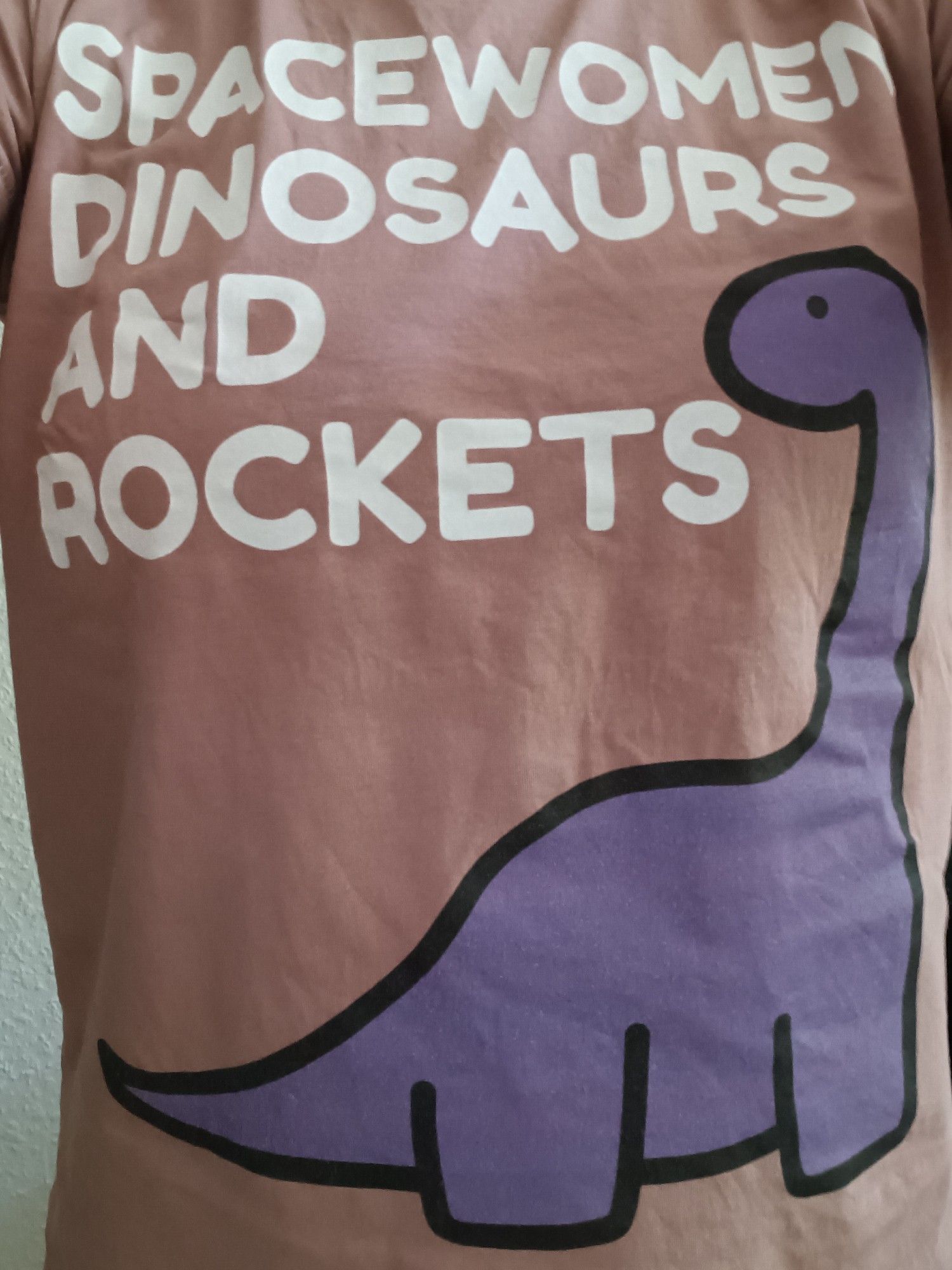 Altrosa Shirt mit einem lila Langhals-Saurier und dem weißen Aufdruck "Spacewomen Dinosaurs and Rockets". Es wird getragen, man sieht aber nichts von der Person.