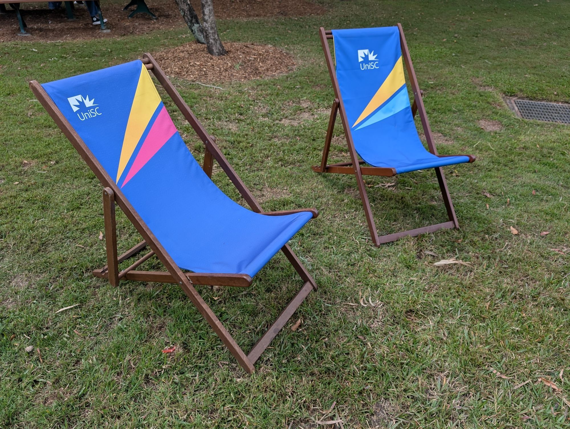 Two blue deck chairs with bright pink and yellow or blue and yellow stripes and a UniSC logo on a grassy patch.