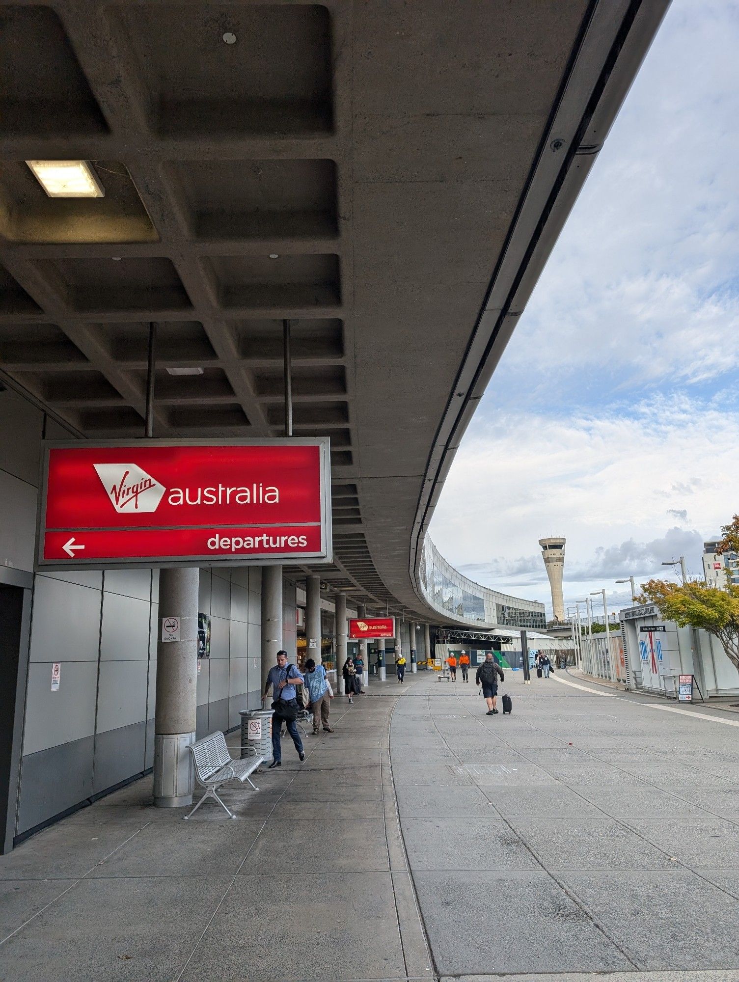 The outside of the Brisbane Domestic Airport terminal with the Virgin Australia red departures sign in view. The gentle curve of the building arcs away with the air control tower at its end, clearly visible against an overcast sky.