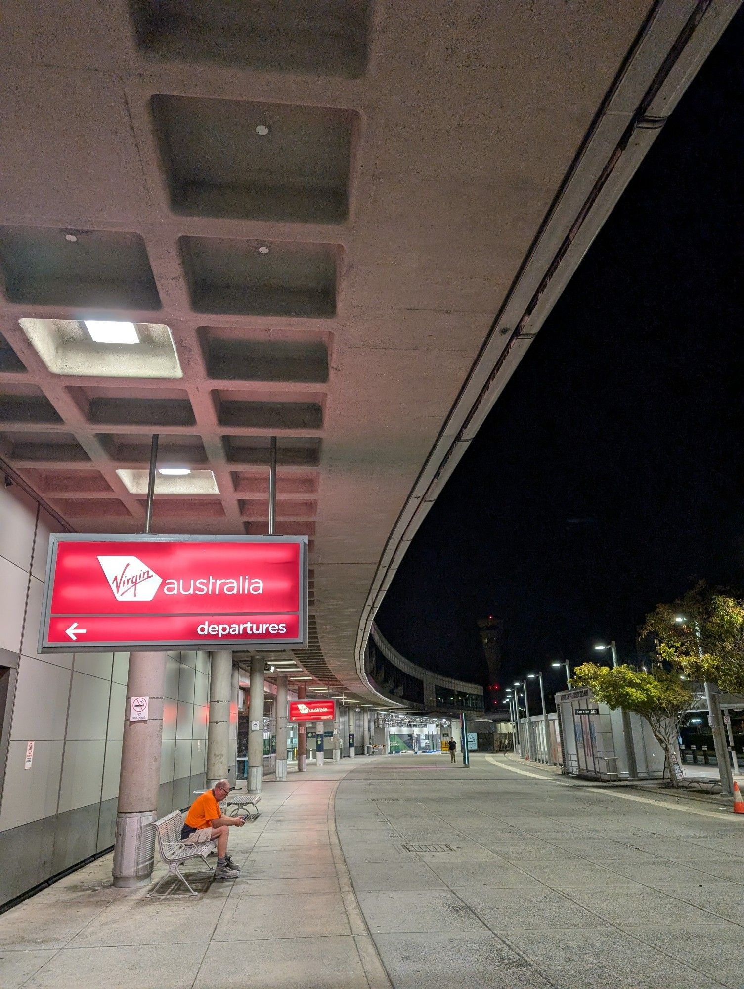 The outside of the Brisbane Domestic Airport terminal with the Virgin Australia red departures sign in view. The gentle curve of the building arcs away with the air control tower at its end, barely clearly visible against the black pre-dawn sky.