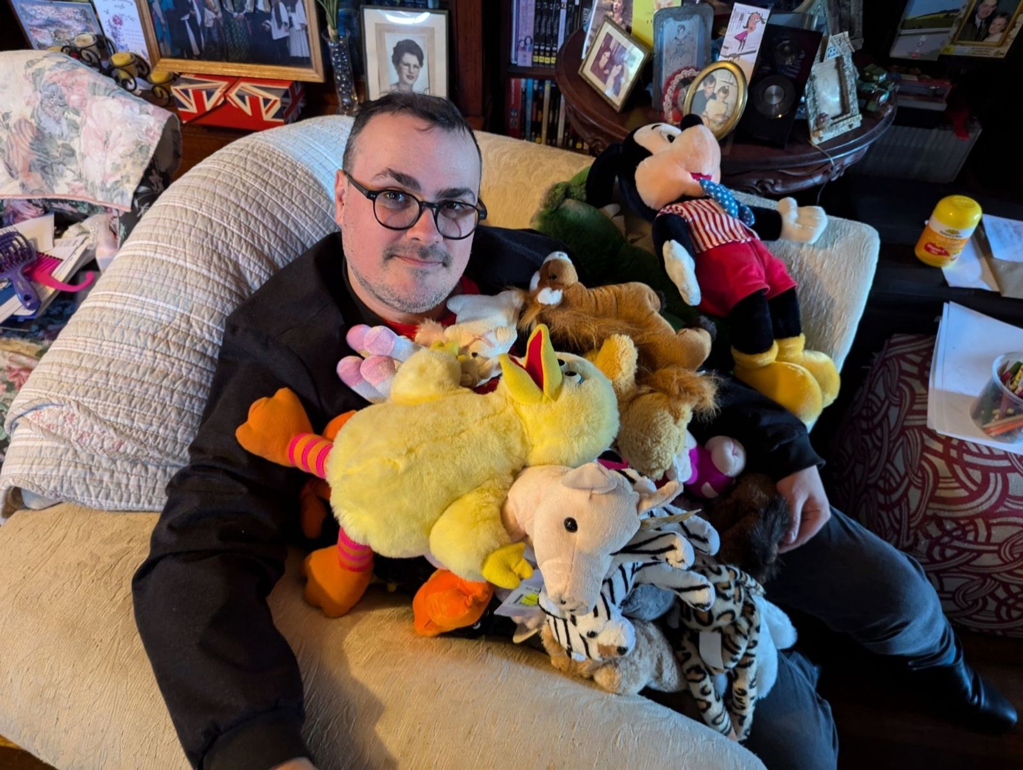 A middle aged man sits on an arm chair, covered in stuffed toys, including Micky Mouse, Big Bird, a unicorn and more.