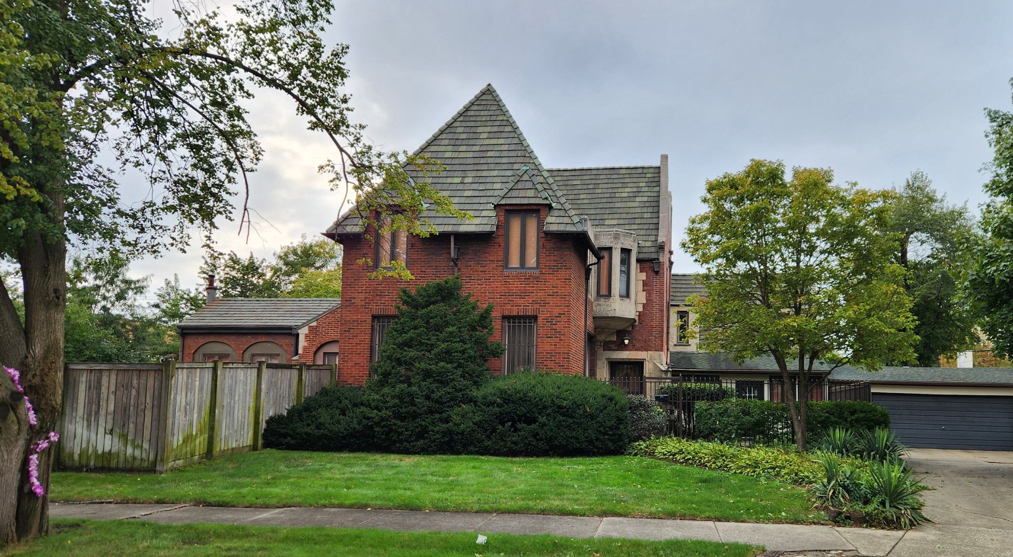 A photo of a large two-story Tudor Revival style home, set back significantly from the street and partially obscured by tall hedges and a tall wooden fence, taking up multiple adjacent city-size lots. The house features a handsome tall slate roof, a small stone turret on the inner part of a corner over its front door, and a flat-roofed midcentury garage off to one side.