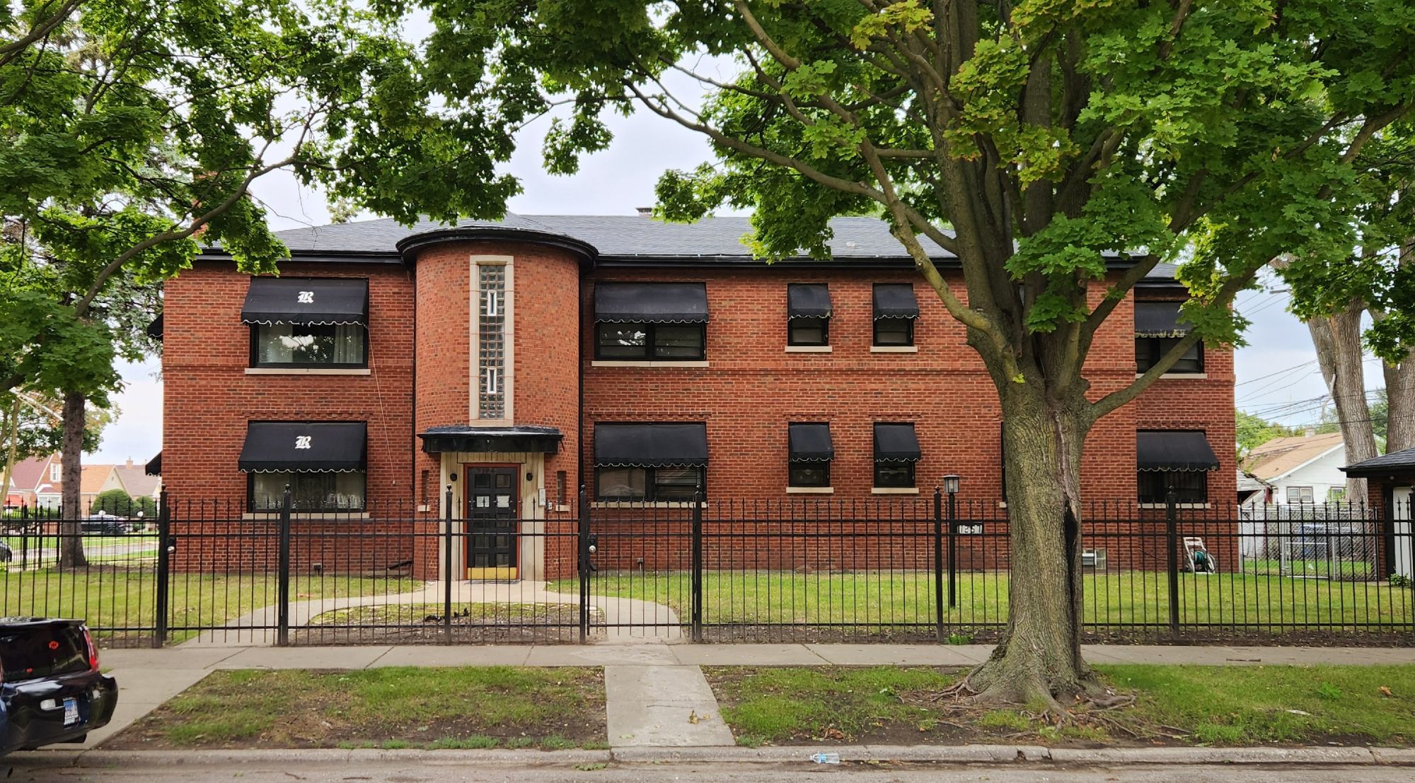 A wide two story red brick house sits on a double lot. Its style is between Art Deco and Art Moderne, with some accented horizontal brick lines on the facade providing massing and cloth awnings over its many small windows, but the main entrance at the end of a U-shaped walkway is a curved cruise ship-like tower with a small vertical window made from glass blocks and surrounded by smooth stone above the door.