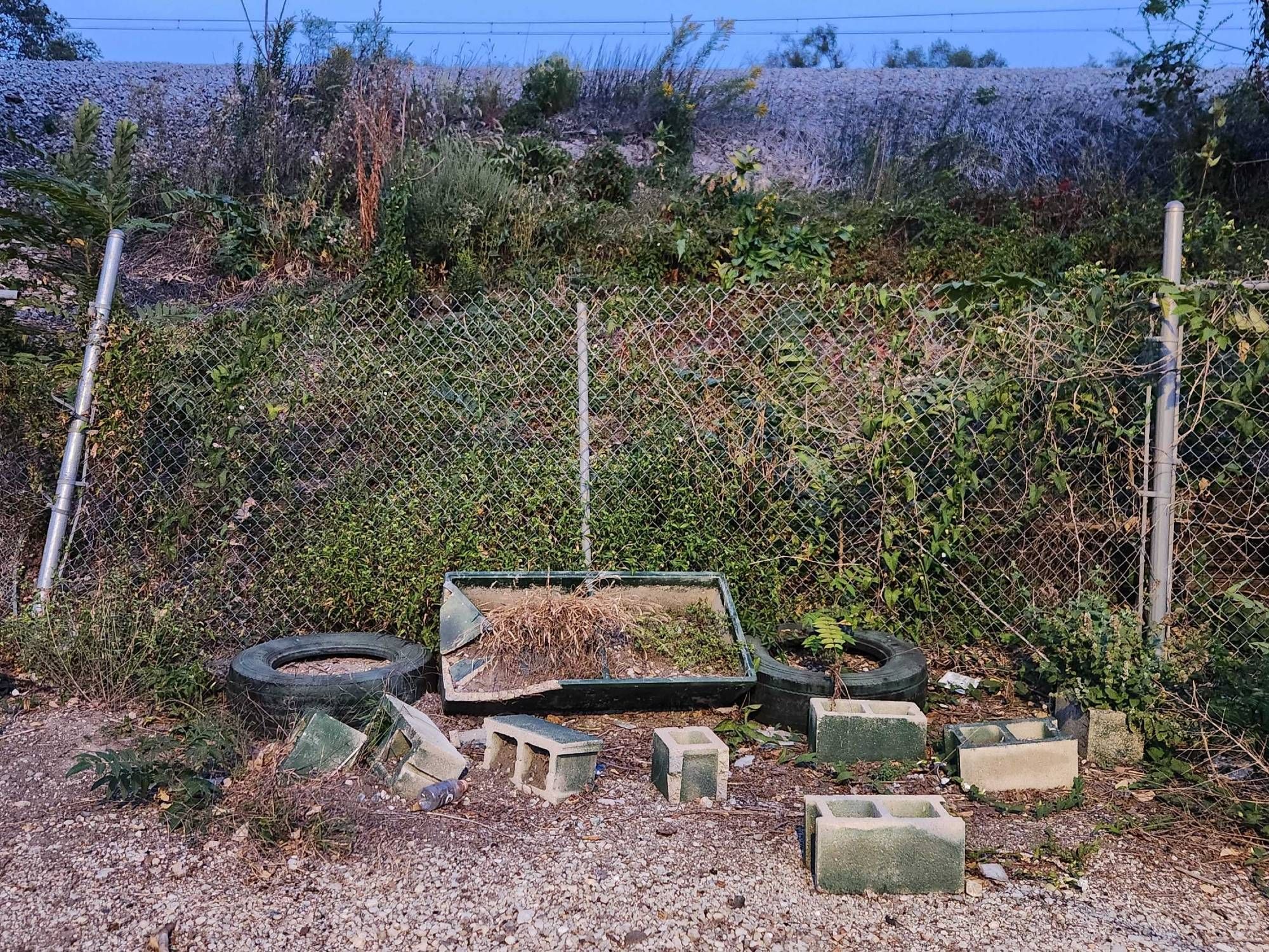 A photo of cinderblocks, tires, and an empty flower box strewn about on gravel in front of a fenced railway embankment