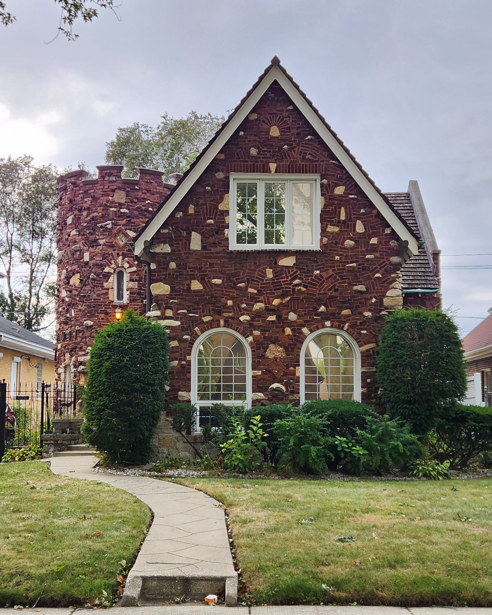 A photo of a Tudor revival style brick home with a very elaborate rusticated brick facde, interspersed with gray stones of varying sizes. The facade's primary brick covering is not a series of even rows; instead, it's a carefully assembled layout of bricks of varying size and shape making up small curves, patterns, and so on, like a fieldstone wall, but made of brick. The house features a castle-like circular turret along the side, and tall front windows with curved tops.