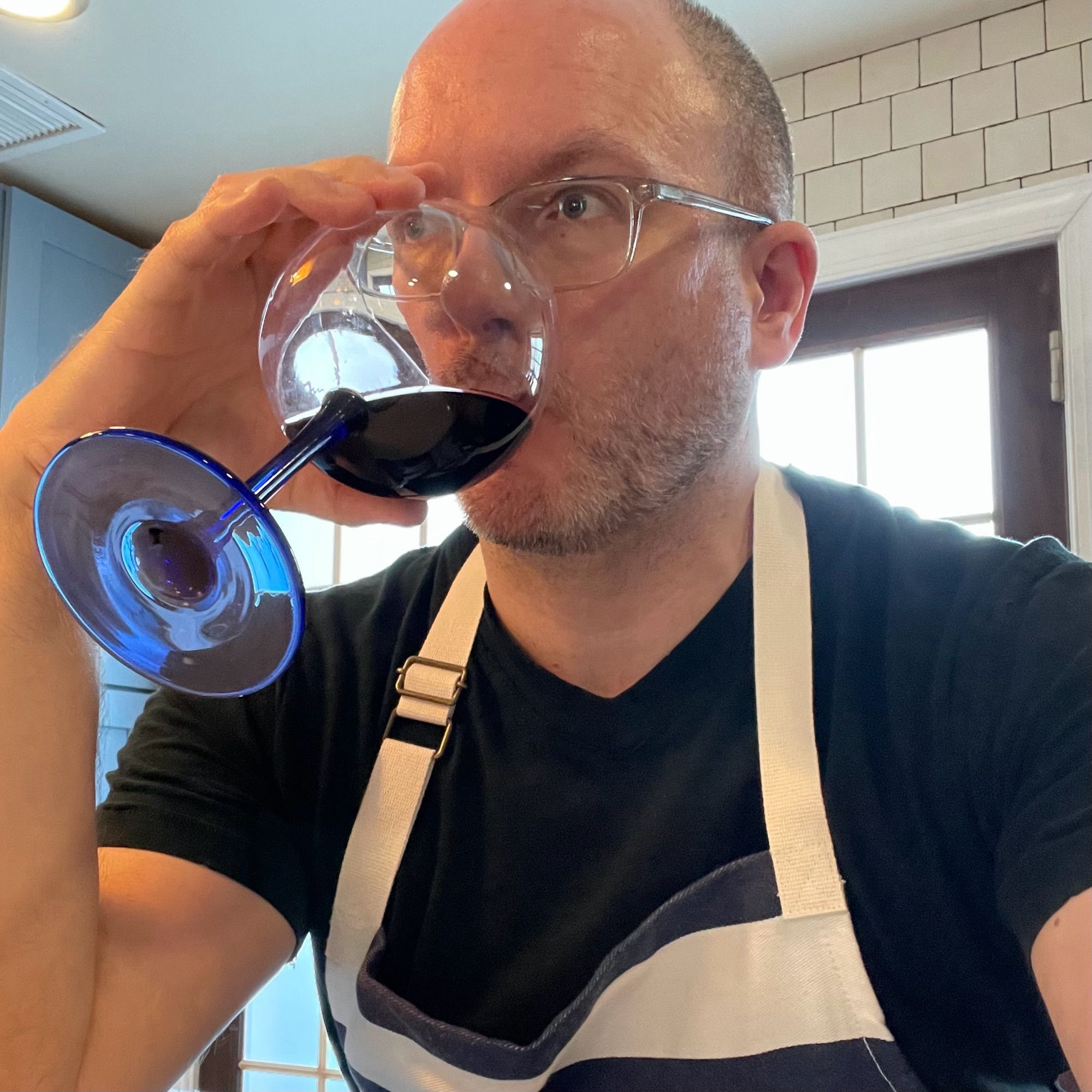 A picture of me wearing an apron sitting in my kitchen drinking a glass of red wine.