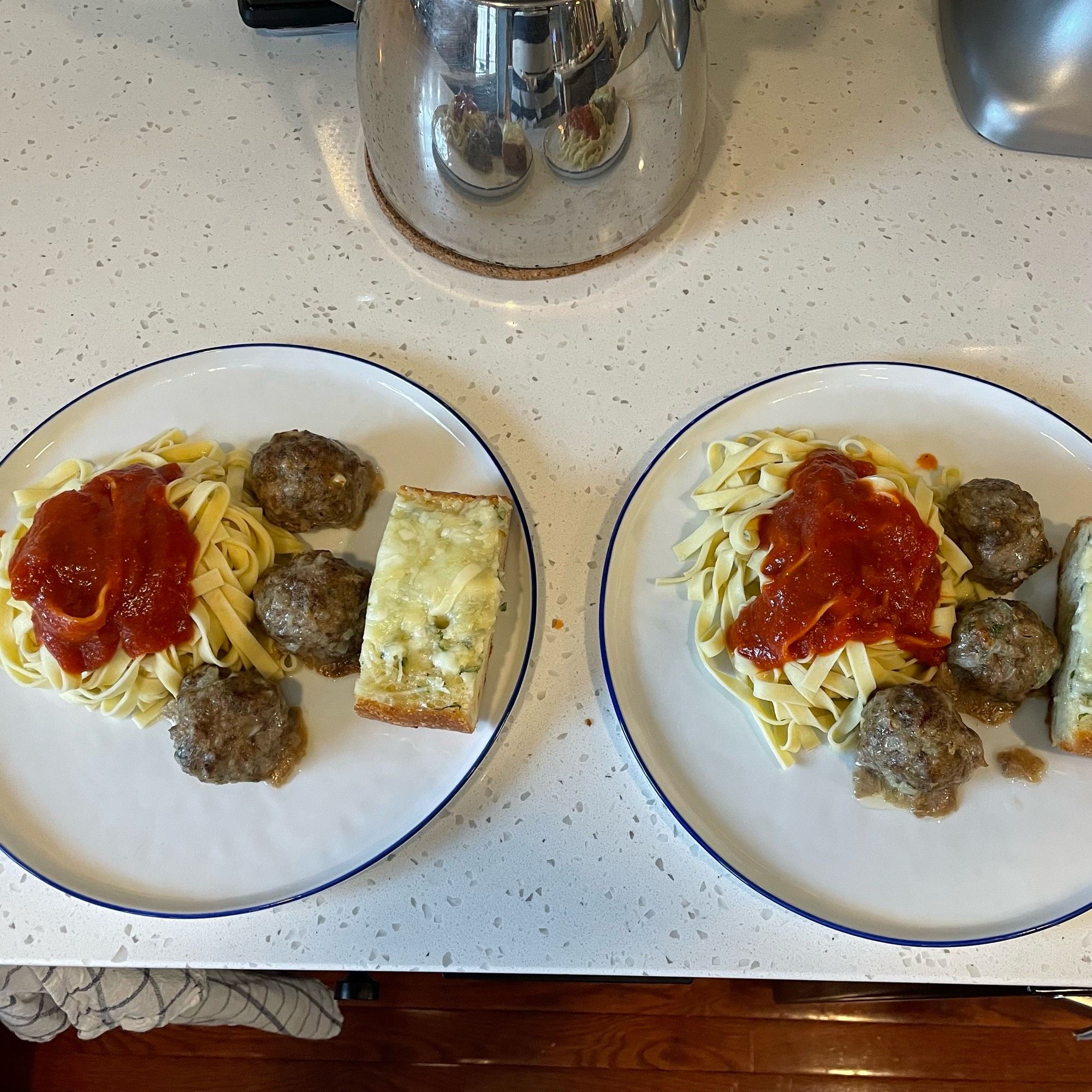 Two plates sit on a counter. Both have a nest of fettuccini topped with sauce, and to the side are three meatballs each and a slice of freshly baked garlic bread.