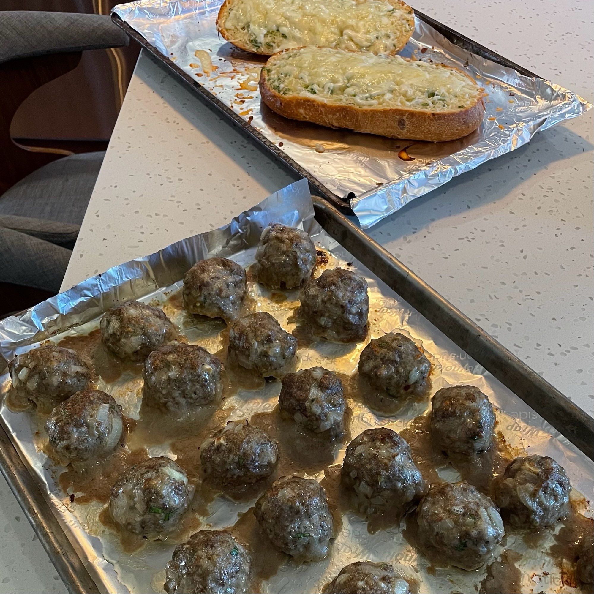 Two trays are on a counter. One has freshly baked garlic bread. The other has rows of freshly baked meatballs.