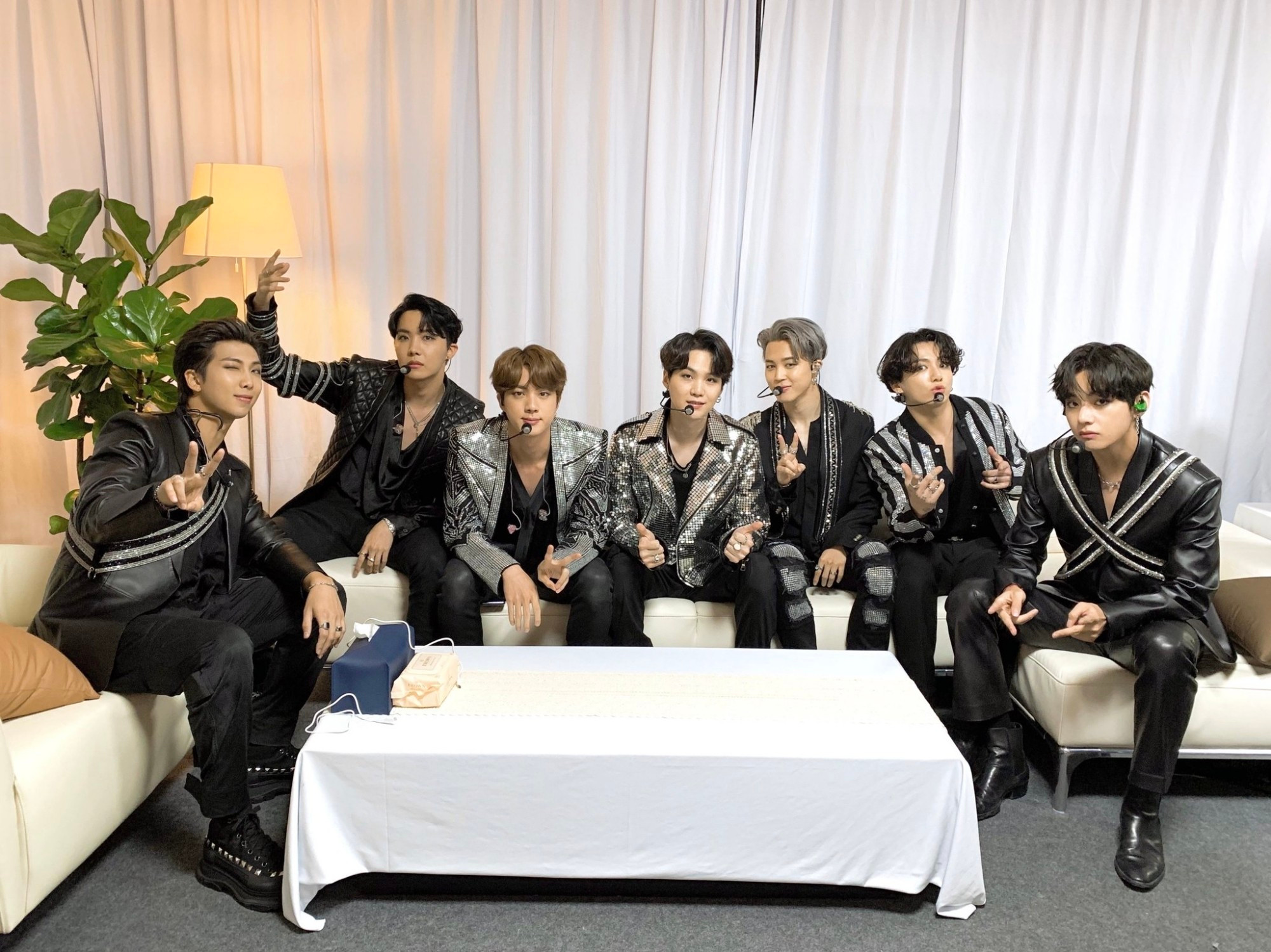 It's a group photo of BTS sitting on a couch in a room, dressed in black and shiny jackets/shiny adorned jackets.