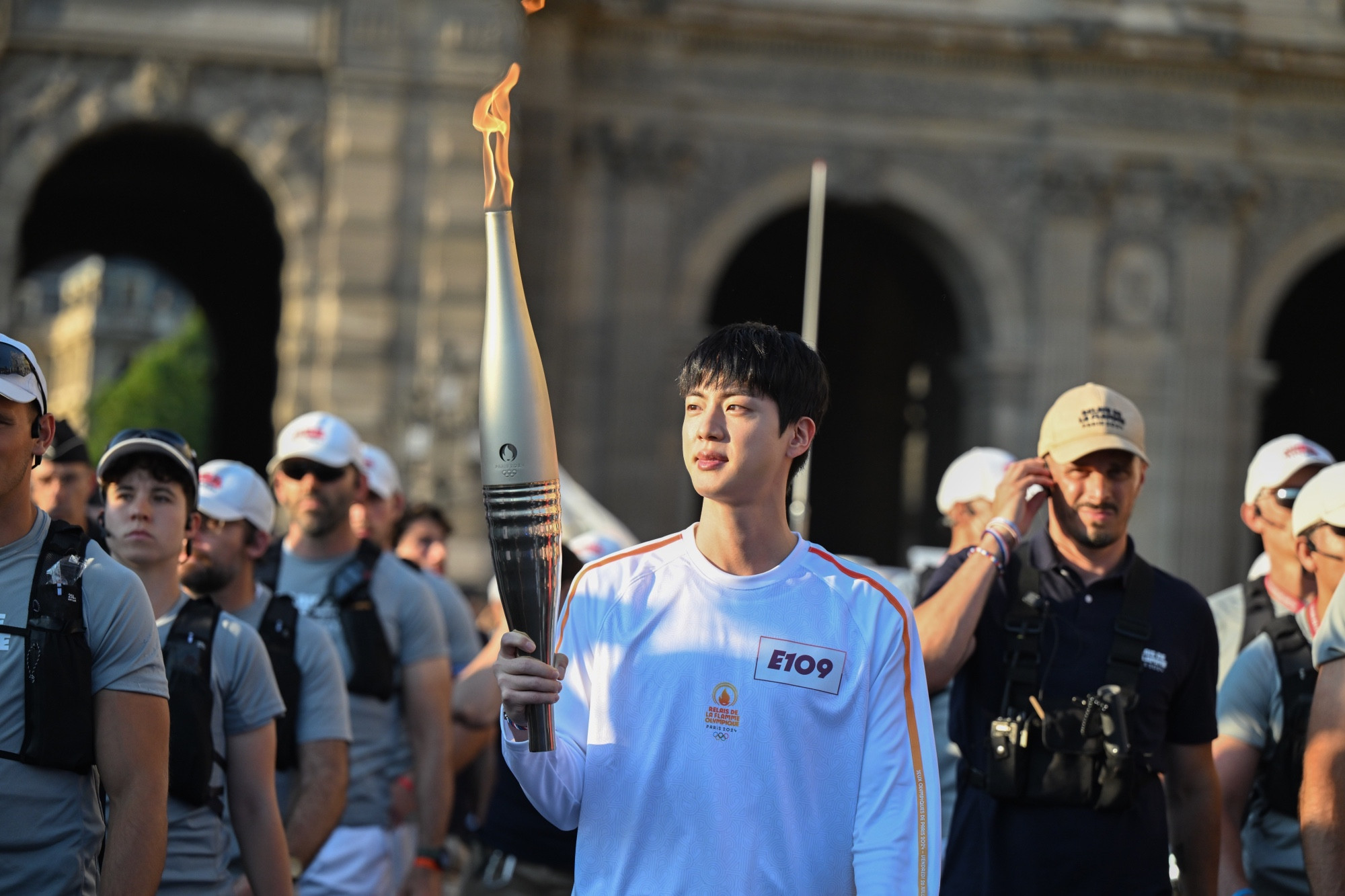 Jin in white Olympics uniform at the torch relay holding the torch while surrounded by security.