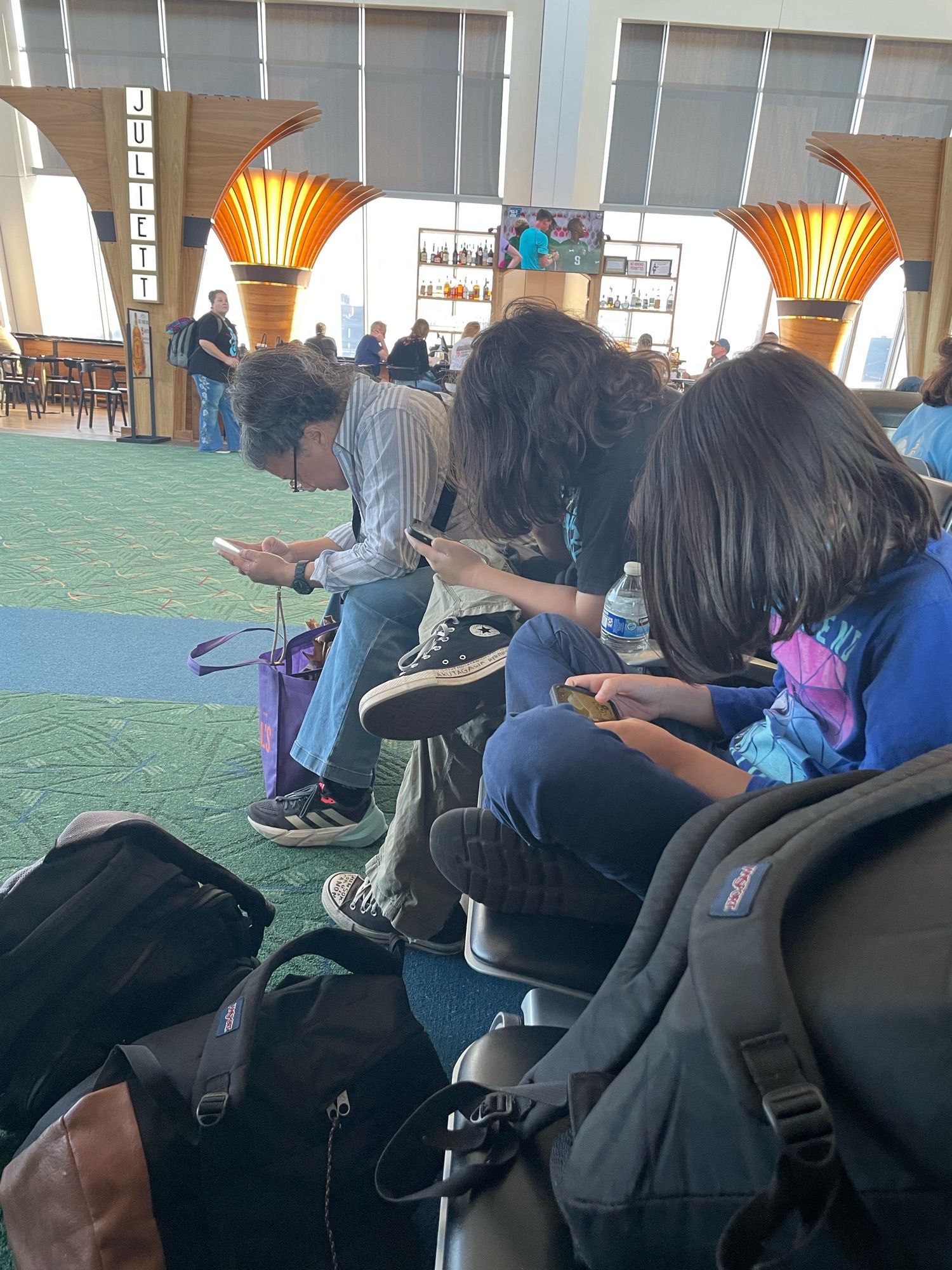 3 people sit in an airline boarding area, their heads pointed down, looking at their phones, in a symmetrical way.