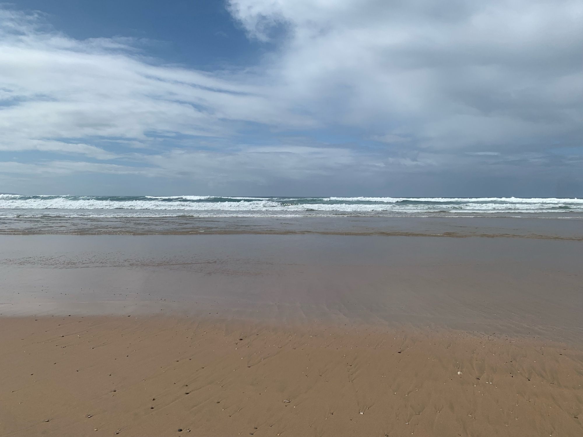 Photograph of the shoreline along a sandy beach, looking out into the vast Southern Ocean. There are a few ominous gray clouds in the far distance