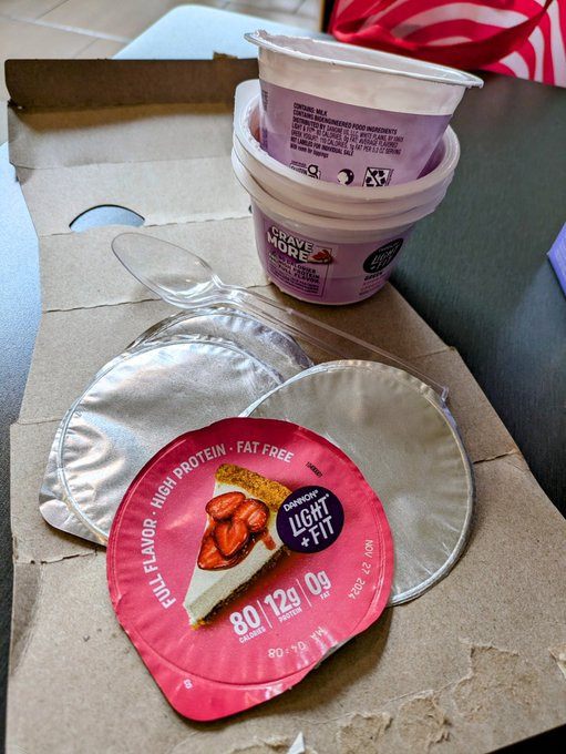 Four empty yogurt cups stacked one inside another, with the topmost one cantilevered at a "jaunty" angle. Directly in front of this tower of yogurt cups rests a single plastic spoon, and in front of this spoon are the four peeled off foil lids-- one which proclaims "FULL FLAVOR * HIGH PROTEIN * FAT FREE' in white lettering above a graphic of a strawberry cheesecake slice. All of this is resting on a piece of cardboard, which turns out to have been the packaging for these four yogurt cups, torn open and converted into an impromptu placement