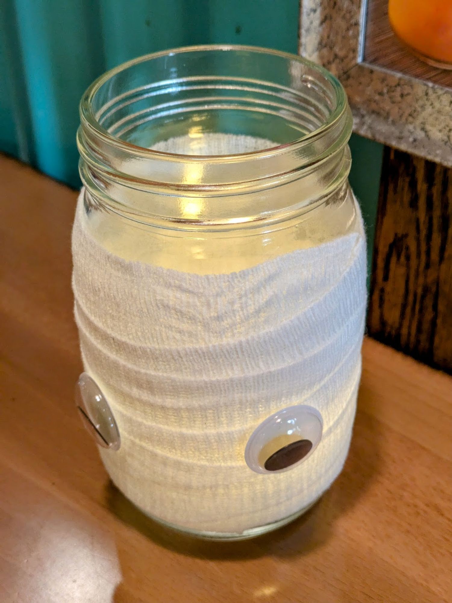 A lit candle, inside of a glass jar, which has been wrapped in a thin gauze-like material. Two googley eyes have been glued to the gauze, creating the appearance of a cute ghost-like "creature" on a restaurant table.