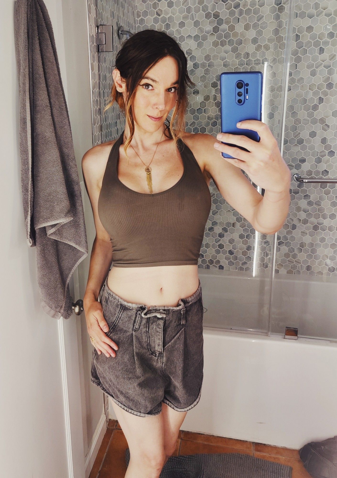 Cara posing for a mirror selfie holding a blue phone and wearing grey denim shorts and a green low cut crop top 