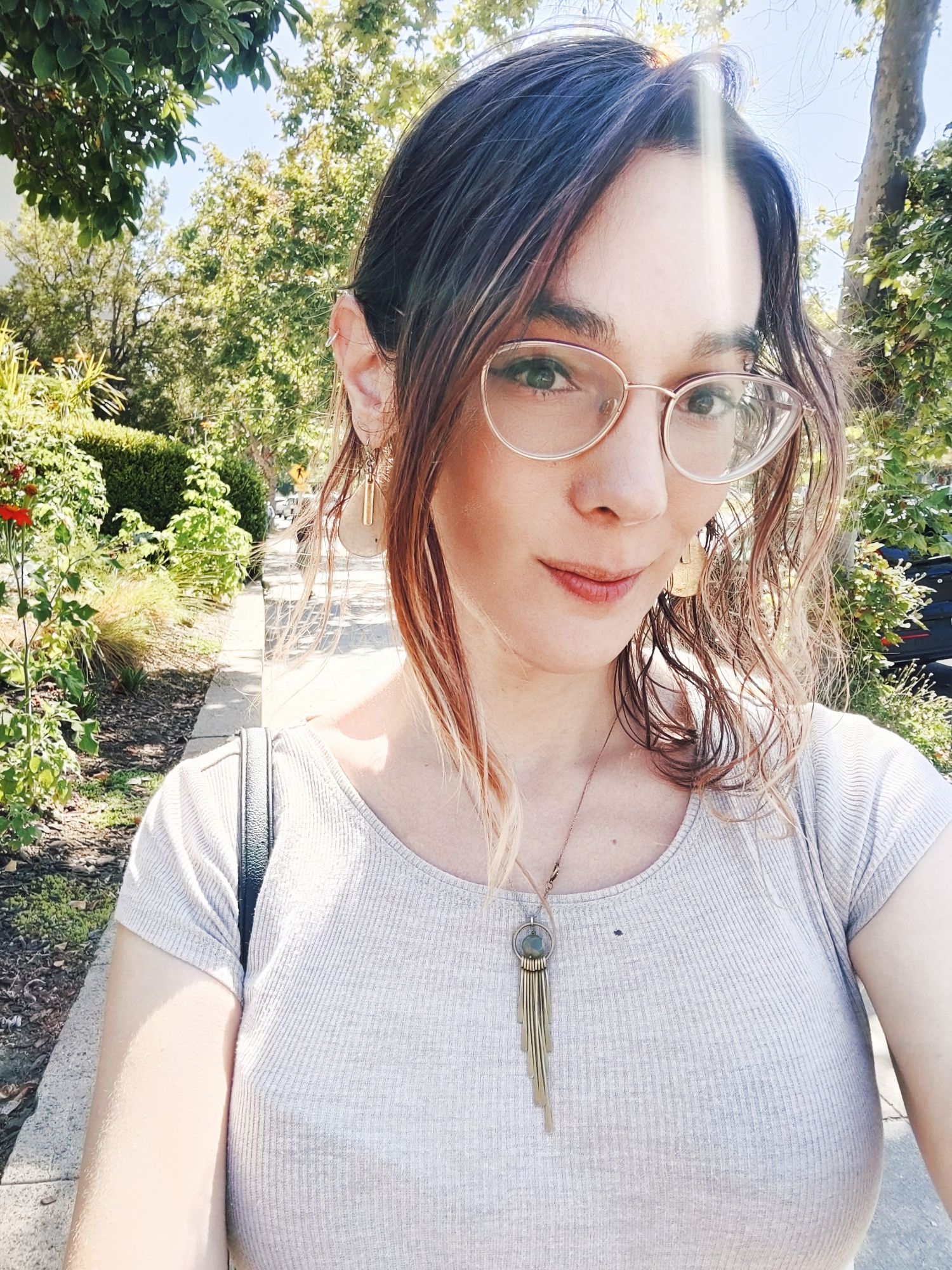 Cara in a tan top and gold glasses, surrounded by trees, half smiling at the camera 