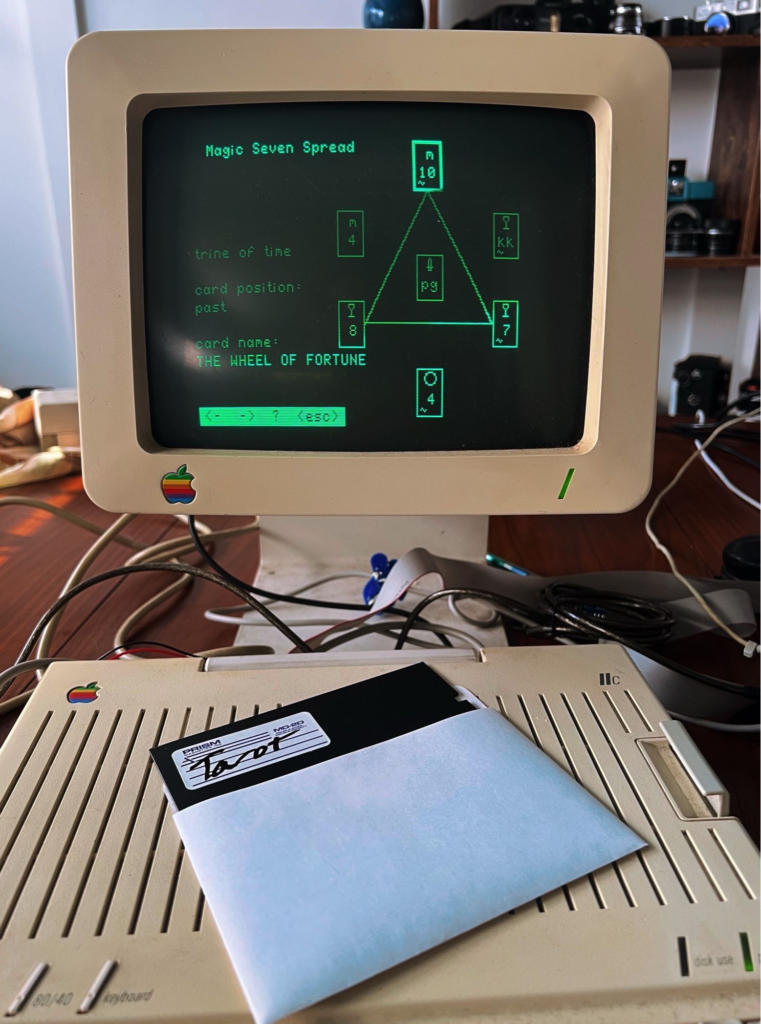 An old green display and an old Apple IIc computer below it. On top of the computer is a disk labeled tarot, the computer has a seven card tarot spreadsheet in crude graphics and the UI has a wheel of fortune card centered