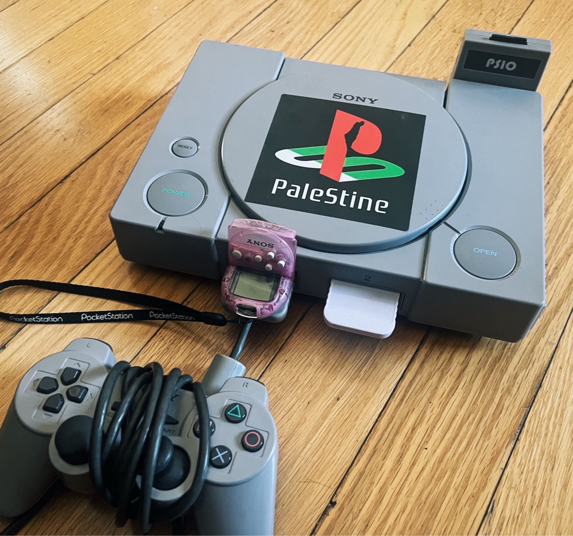 Ps1 with pink pocket station and dual shock controller, and a large sticker that reads “Palestine” in the PlayStation font, with the Ps logo done up in Palestinian flag colors with an image of Palestine in the center of the P