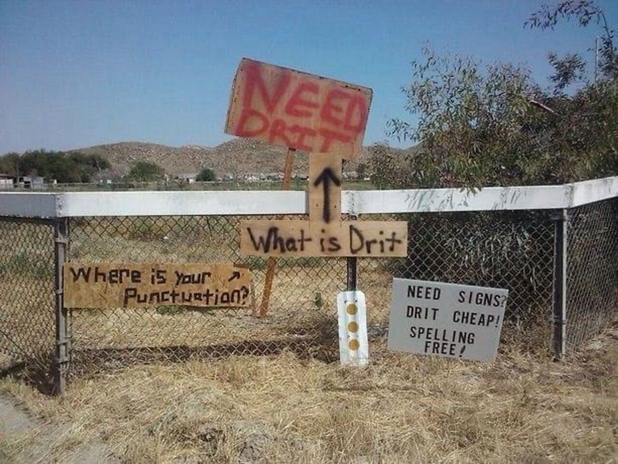 A rustic handmade sign that says “NEED DRIT” and several other signs below it, mocking it. “What is drit?” “Where is your punctuation?” “Need signs? Drit cheap! Spelling free!”