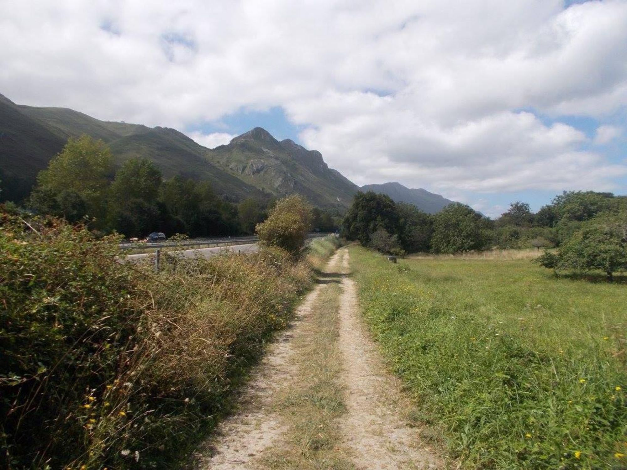 A dirt path surrounded by greenery runs alongside a road, with mountains in the background and a partly cloudy sky.