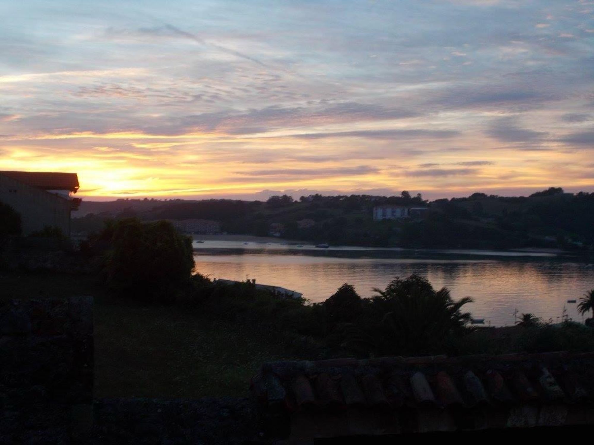 A scenic landscape at sunrise featuring calm water reflecting the vibrant orange and purple hues of the sky. Silhouetted trees and buildings line the shoreline, creating a tranquil atmosphere.