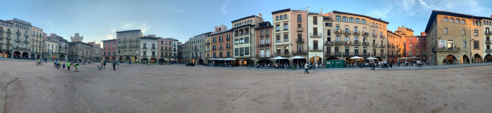 A panoramic view of a lively square surrounded by historic buildings. People, including cyclists and families, enjoy the open space, with some seated at outdoor cafés. The sky features soft clouds, hinting at a pleasant evening.