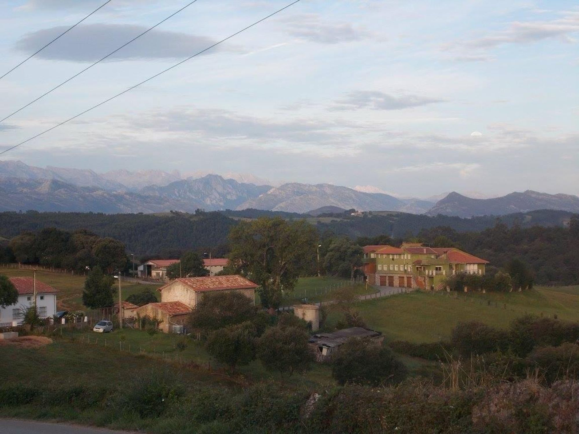 A scenic view of a rural landscape featuring rolling hills, mountains in the background, and several buildings, including houses with red roofs and greenery. The sky is partly cloudy.