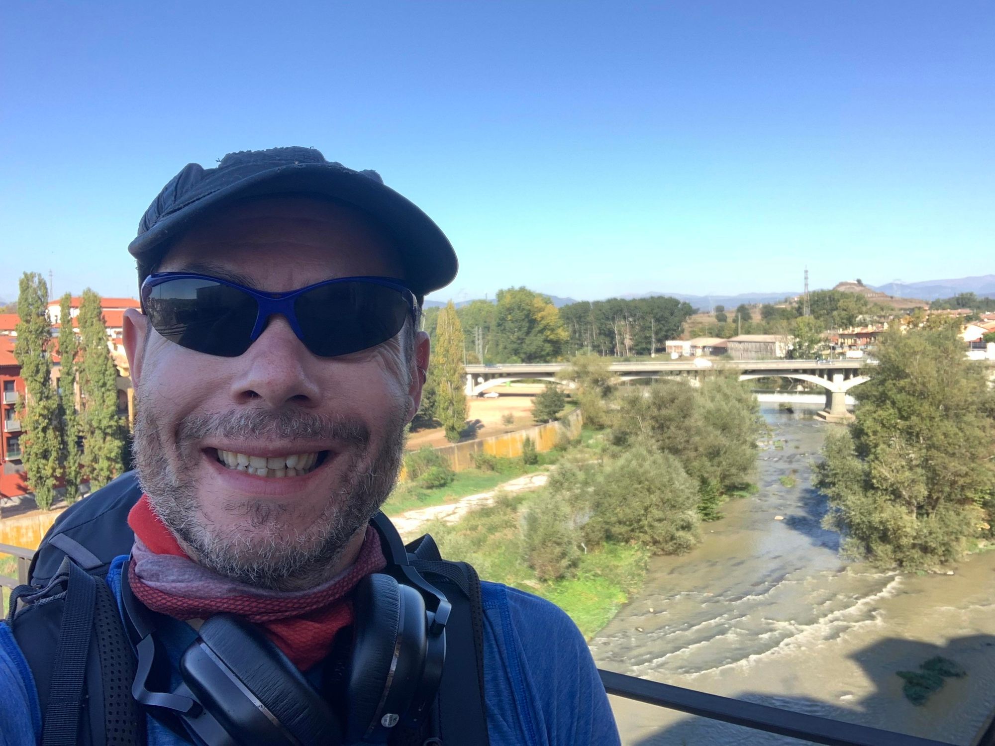 A selfie of me (42 year old cis white gay male in hiker gear) smiling on a bridge with a river and trees in the background. Clear blue sky and hills can be seen in the distance.